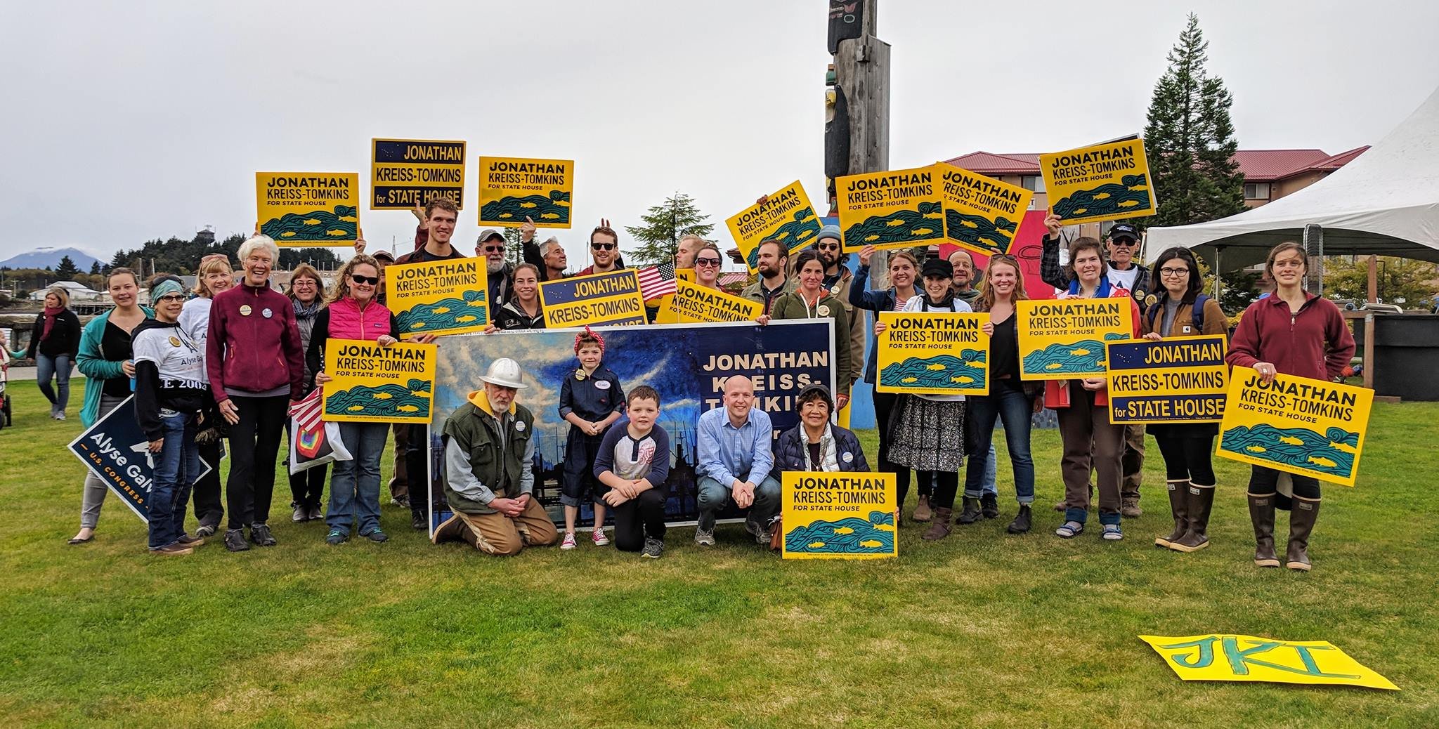 Group of people at a political campaign event holding yellow and blue signs that read "Jonathan Kreiss-Tomkins for State House." Some signs have an illustration of mountains and a river. The group includes adults and children standing on a grassy area with a building, trees, and mountains in the background.