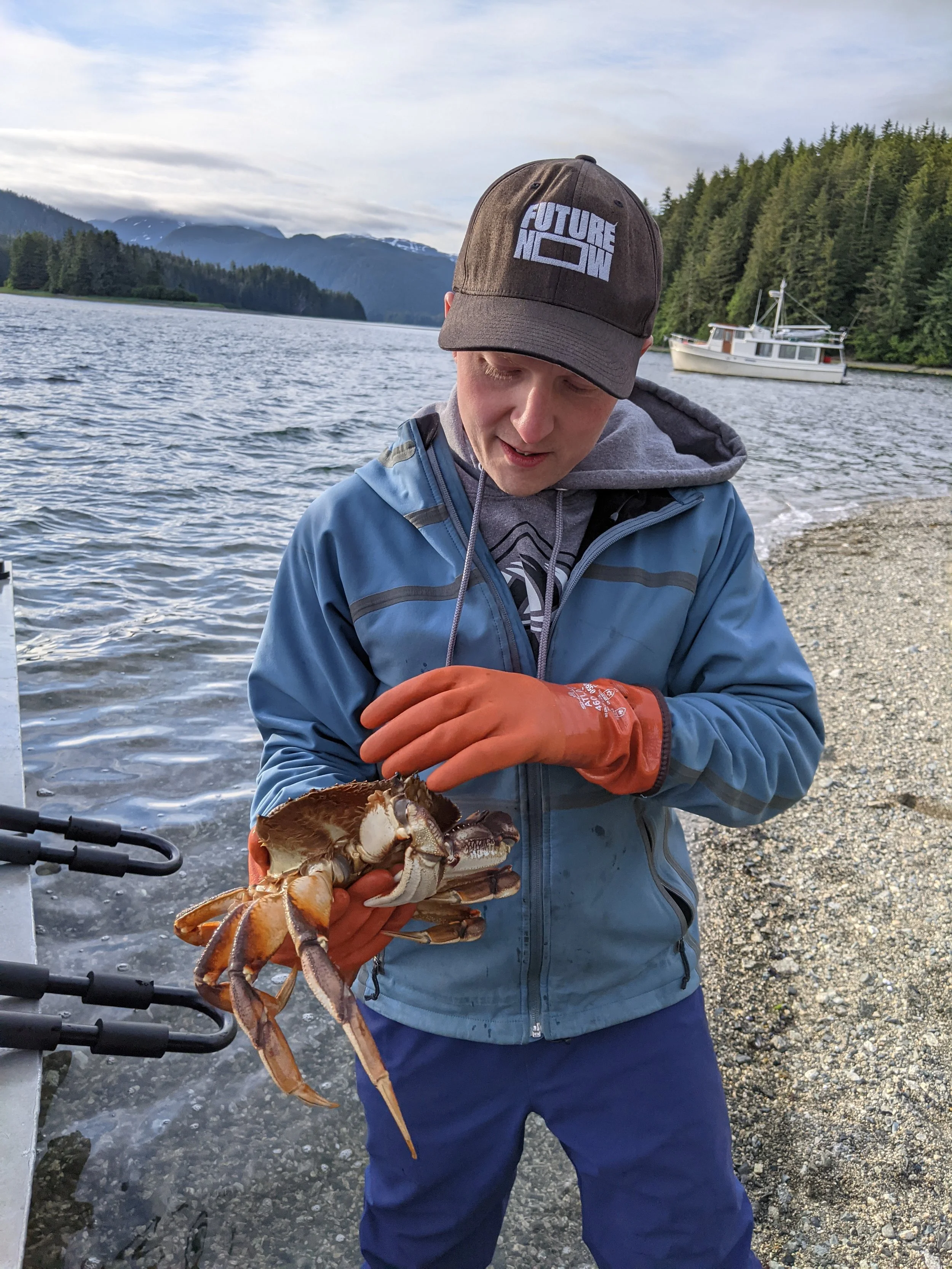 JKT holding a large crab by a rocky lakeshore with boat and forested hills in the background.