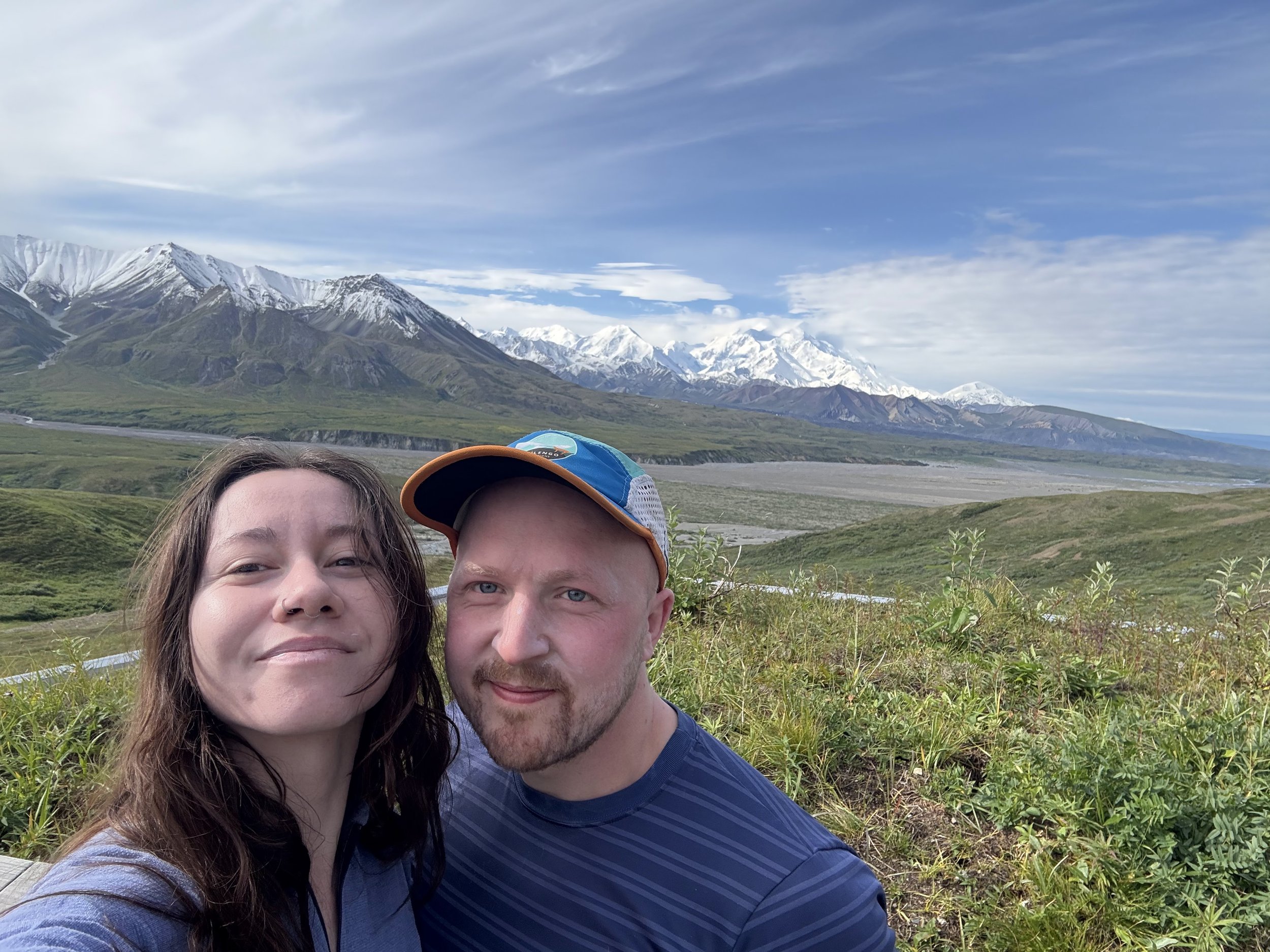 A selfie of a woman and JKT with snow-capped mountains and green hills in the background.