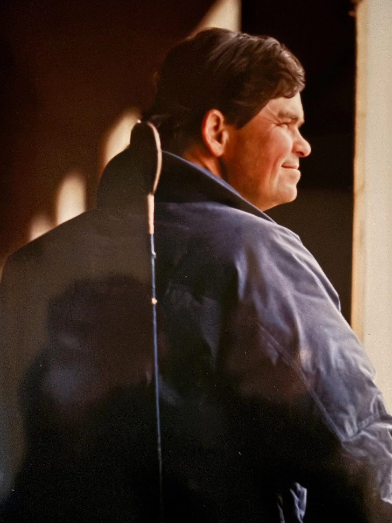 A man with dark hair in a leather jacket, sitting and smiling, looking to the side, in an indoor setting with dim lighting.