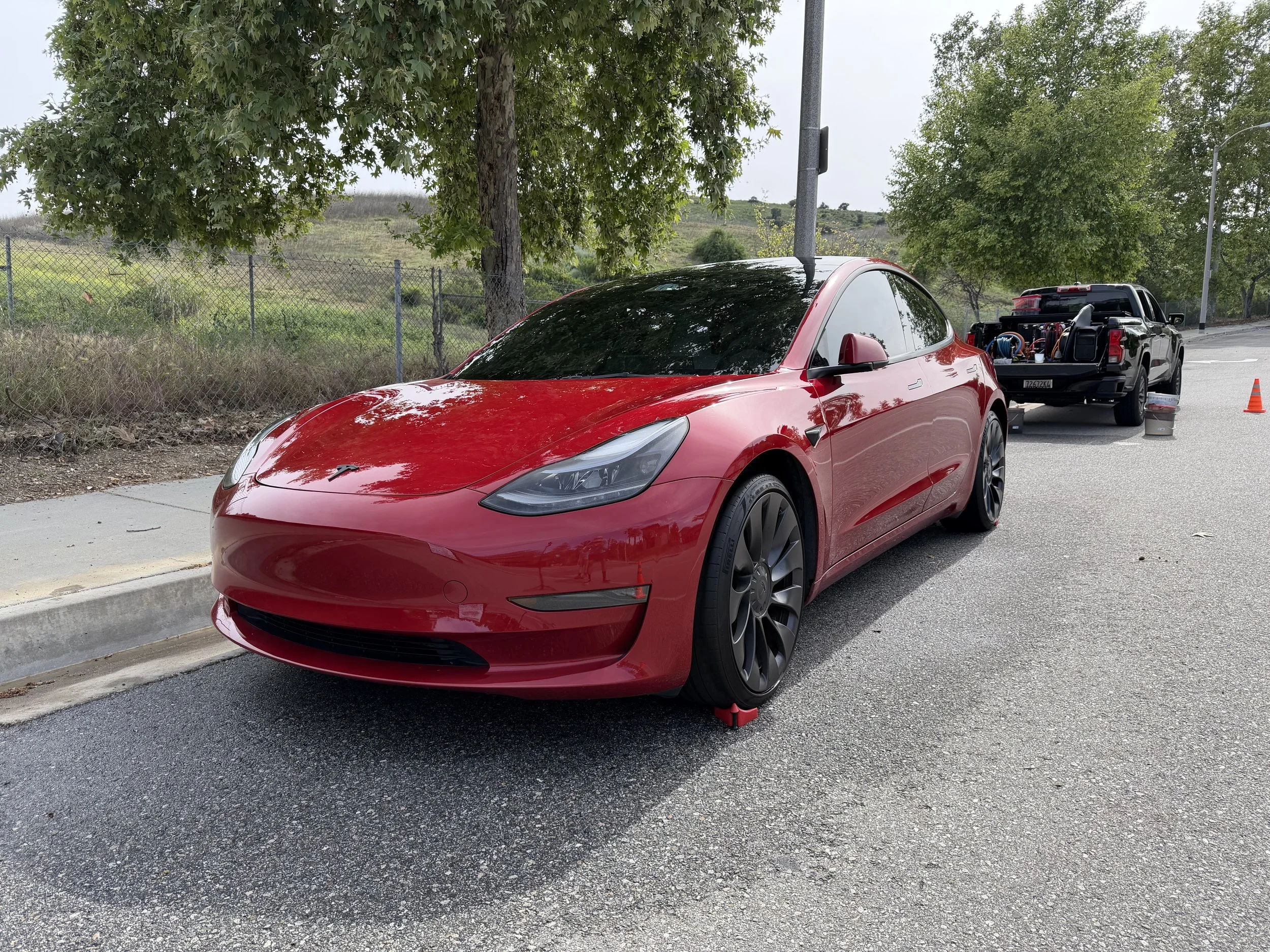 A red Tesla parked on the side of a road with a pickup truck behind it, which has equipment in its bed, orange traffic cones, trees, a fence, and hillside in the background