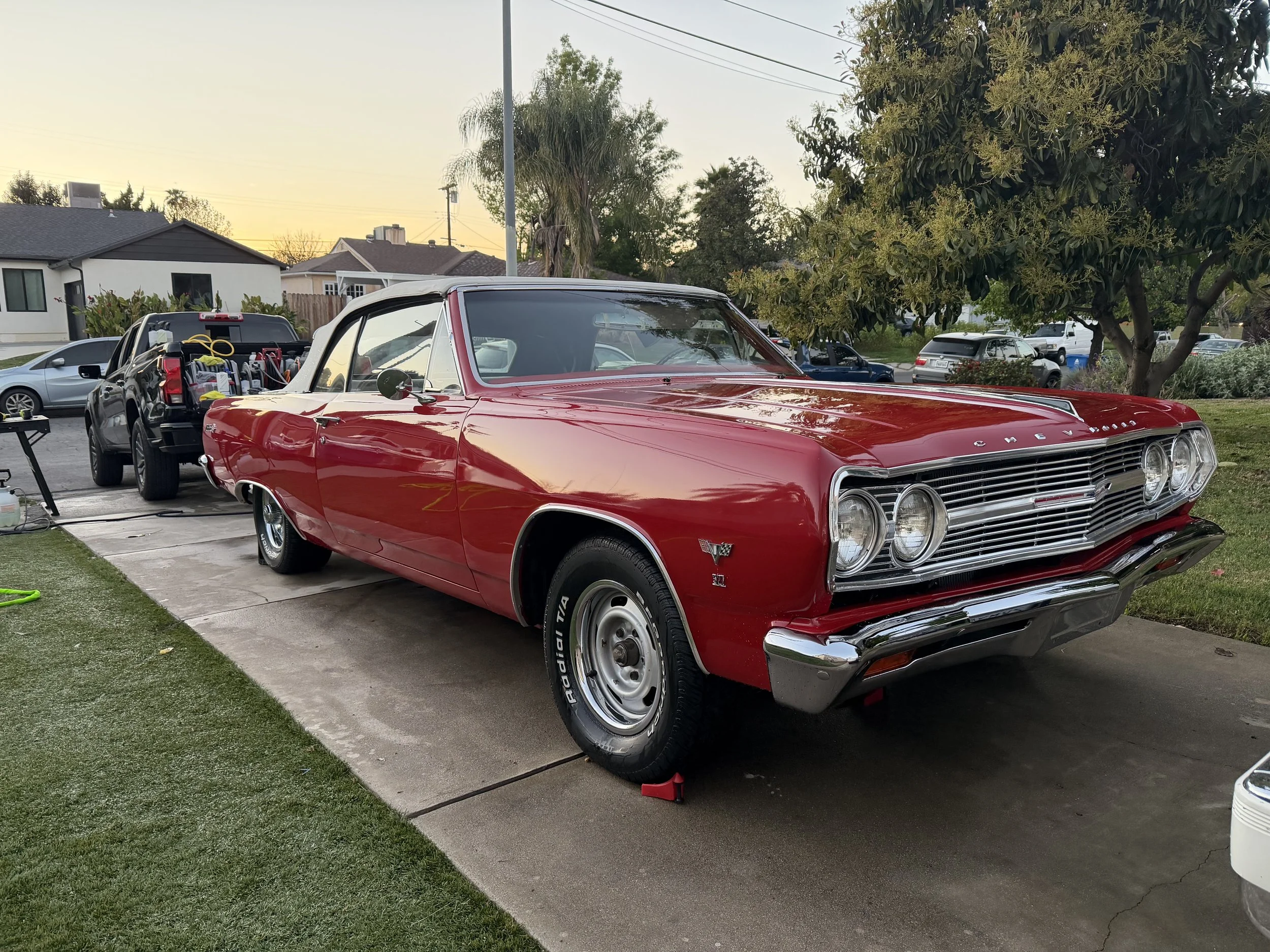 A red vintage Chevrolet car parked on a driveway with a black truck behind it filled with tools and equipment. There are houses, trees, and a sunset sky in the background.