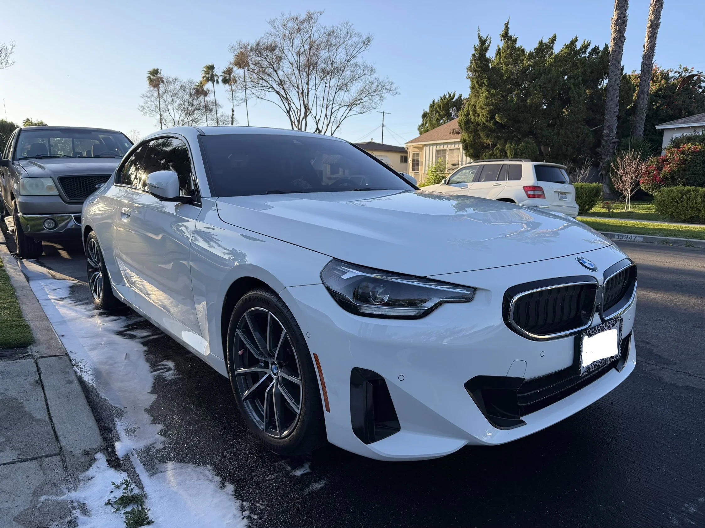 A white BMW car parked on a driveway next to a street with a black SUV parked behind it. There is water on the ground suggesting recent washing or rain, and houses with trees are visible in the background.