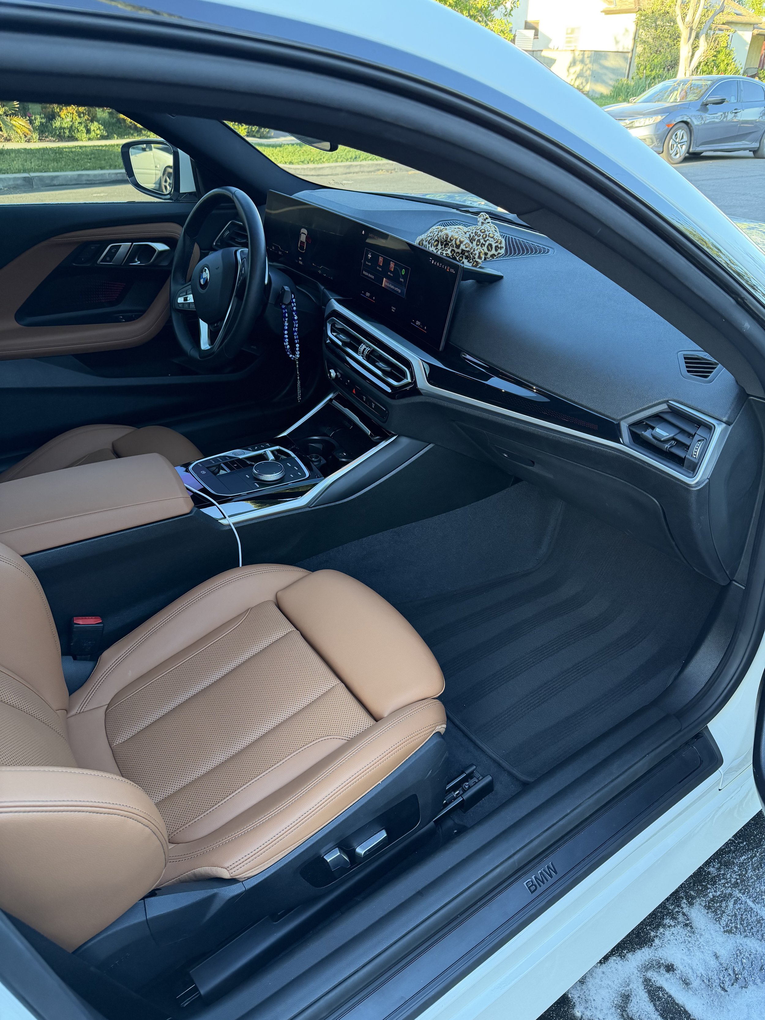 The interior of a BMW car with tan leather seats, a digital dashboard display, and a leopard print stuffed animal on the dashboard. The car is parked outside on a sunny day.
