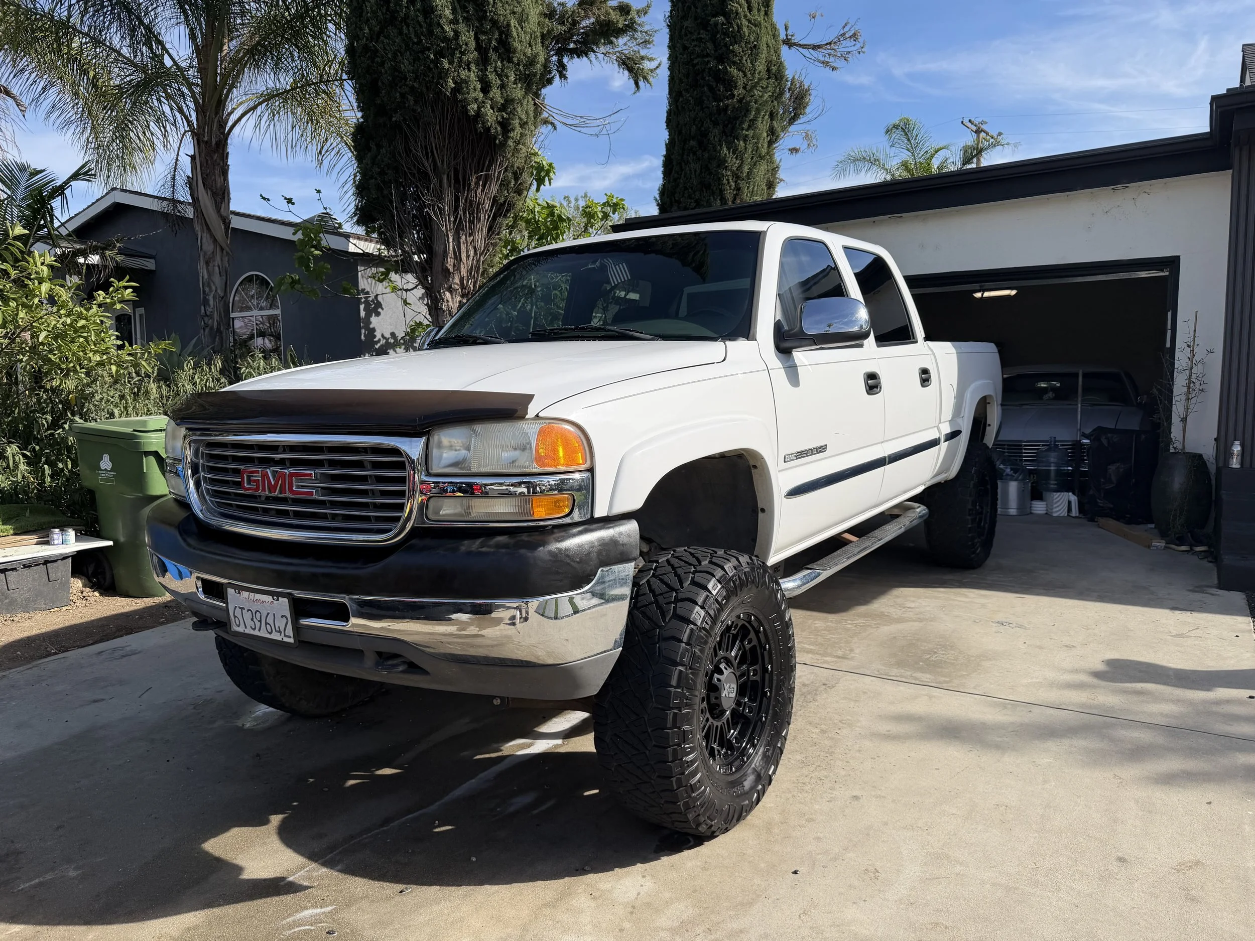 A white GMC pickup truck with large tires parked in front of a garage with the door open. The garage contains a silver vehicle and storage items. There are trees and a blue sky in the background.
