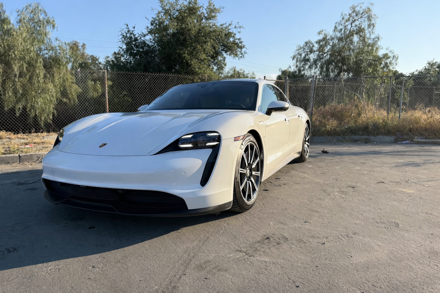 A white Porsche electric car parked on a paved lot with trees and a chain-link fence in the background under a clear blue sky.