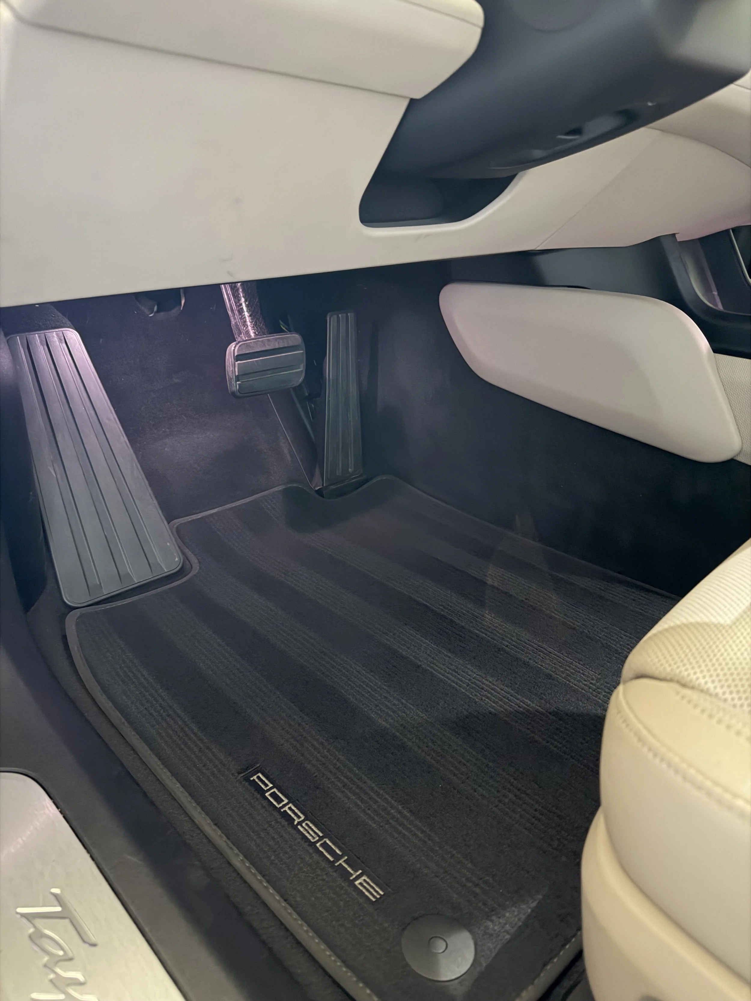 The interior of a Porsche Taycan includes a black floor mat with the Porsche logo, a beige leather seat, and a transparent pedal with a textured grip.