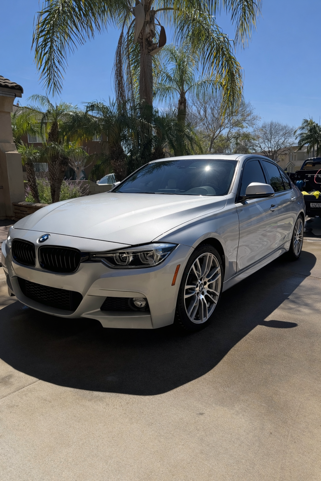 A white BMW sedan parked outdoors in a sunny area with palm trees and a residential neighborhood in the background.