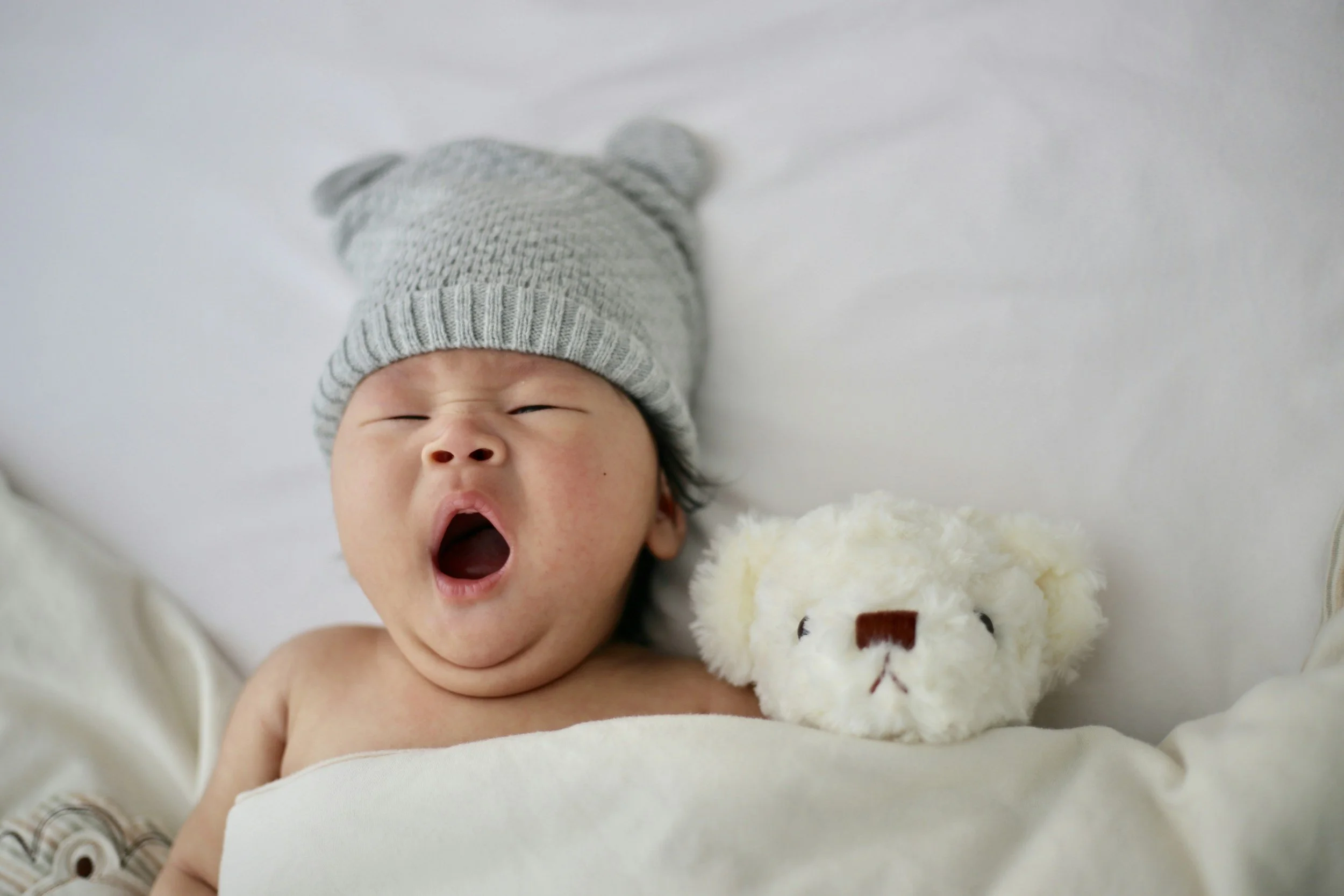 A baby wearing a gray knit hat with bear ears yawning while lying on a white blanket and holding a stuffed animal.
