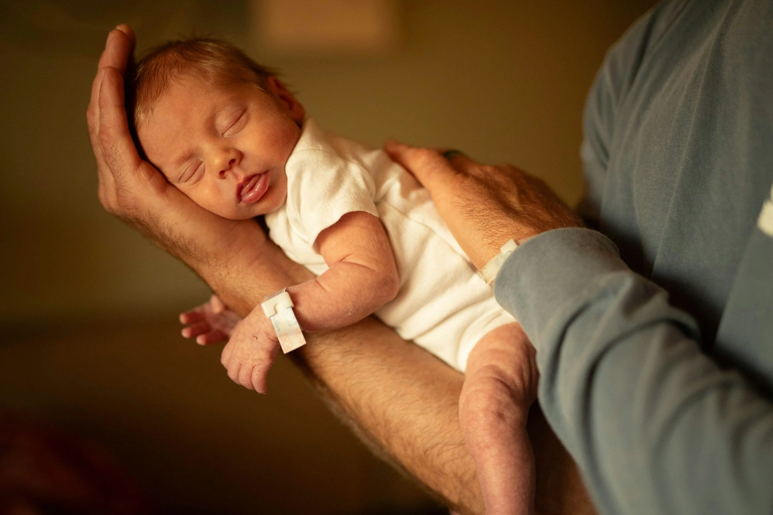 A newborn baby with closed eyes and slightly open mouth, lying on an adult's arm, with the adult holding the baby's head and supporting its body, in a warm environment.