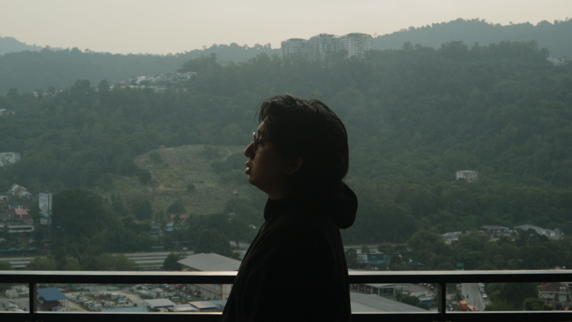 A person with dark hair and sunglasses standing on a balcony overlooking a hilly landscape with buildings and trees.