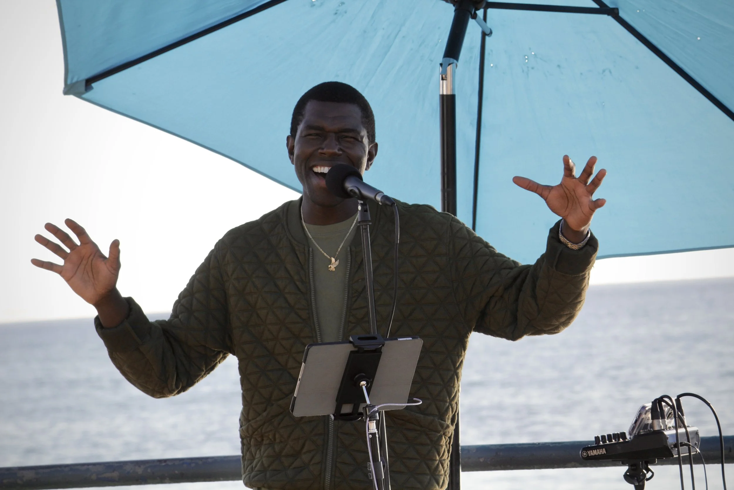 A man singing or speaking into a microphone, smiling with his eyes closed, standing under a blue umbrella outdoors near the water, with a music stand and electronic equipment nearby.