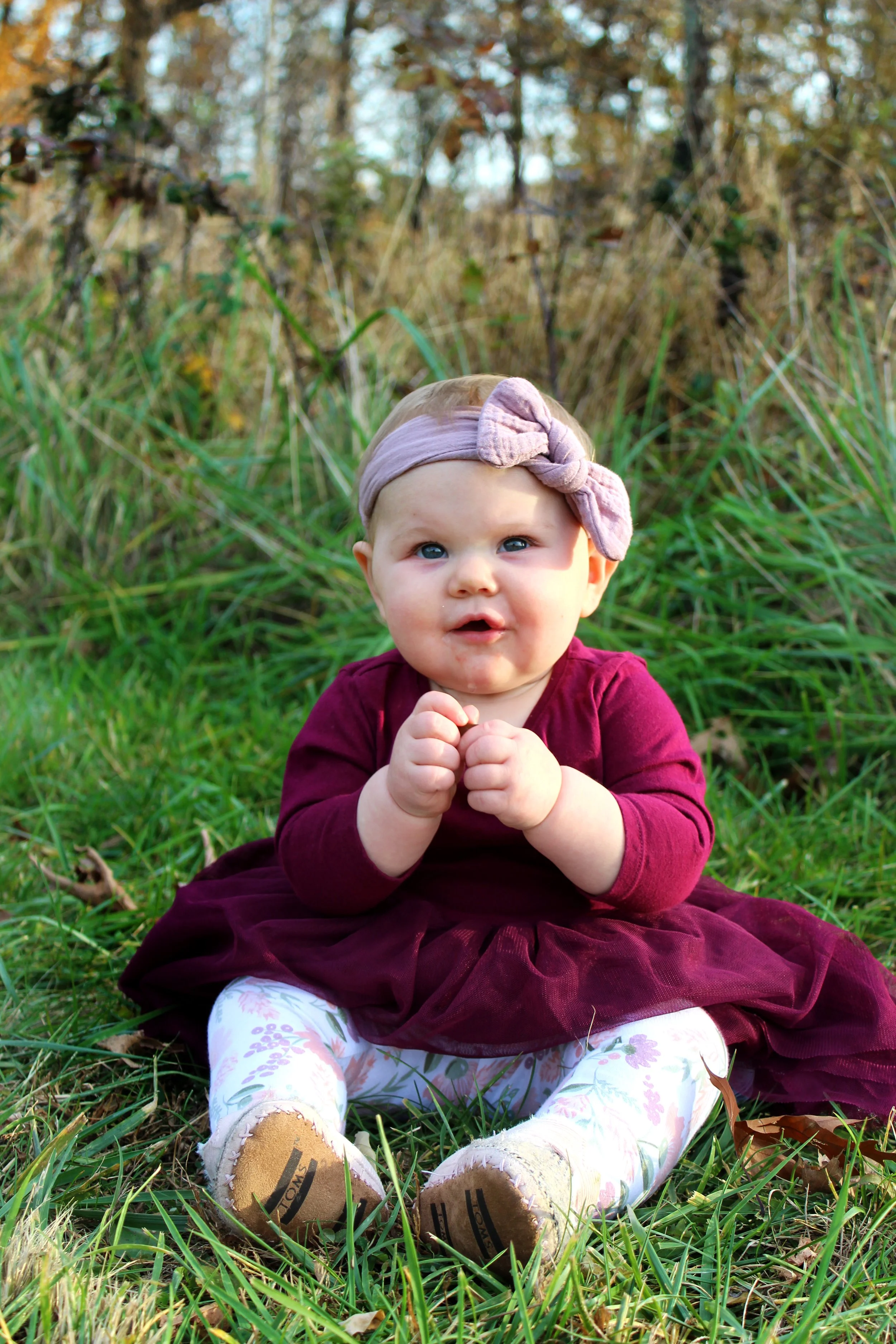 A baby girl sitting on grass in a park with autumn foliage in the background, wearing a purple dress, floral leggings, a pink headband, and tan shoes.