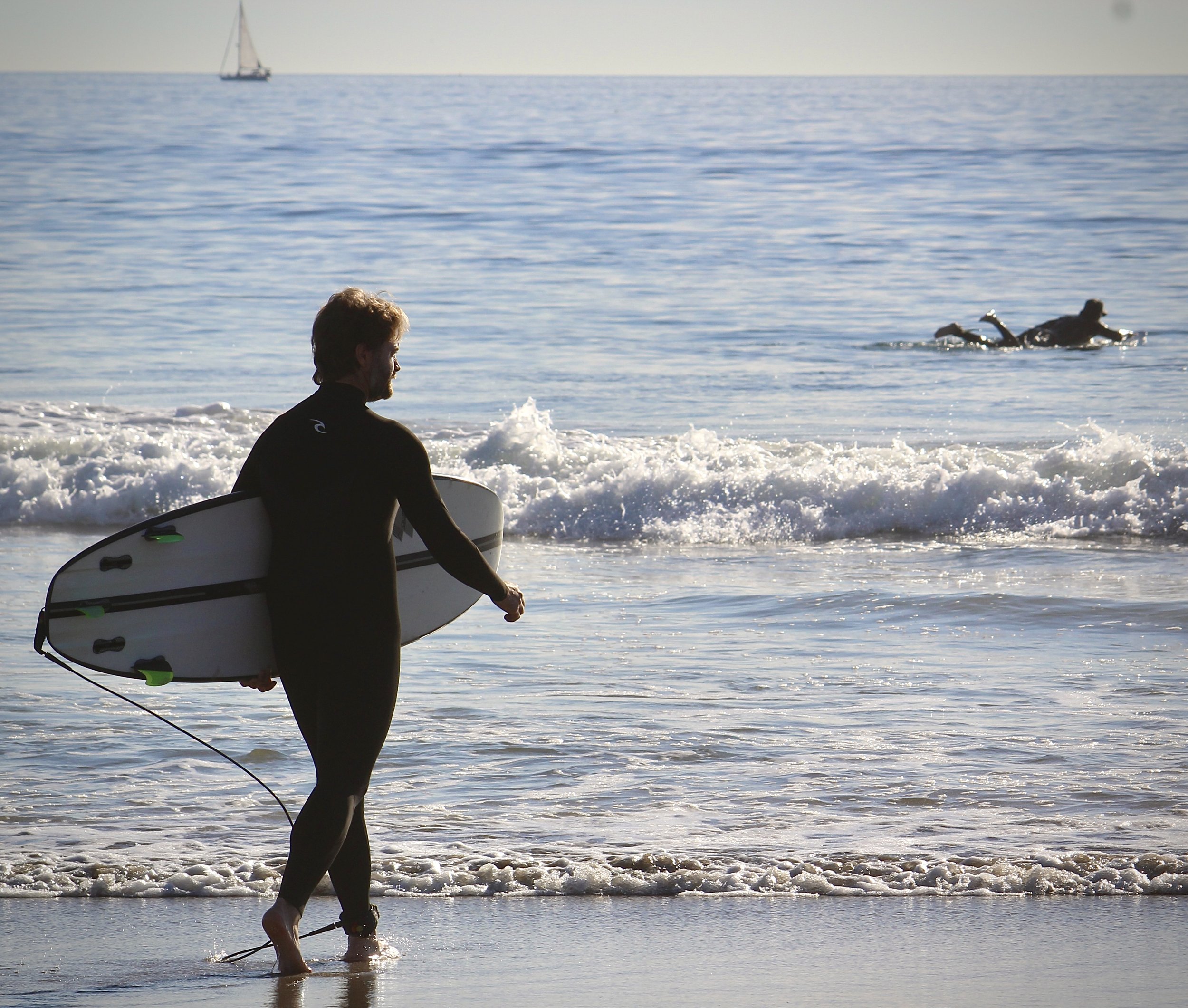 A person with short hair and a beard, wearing a black wetsuit, holding a surfboard, standing in shallow water at the beach while looking out at the ocean. There is a sailboat in the distance on the water and a person swimming nearby.