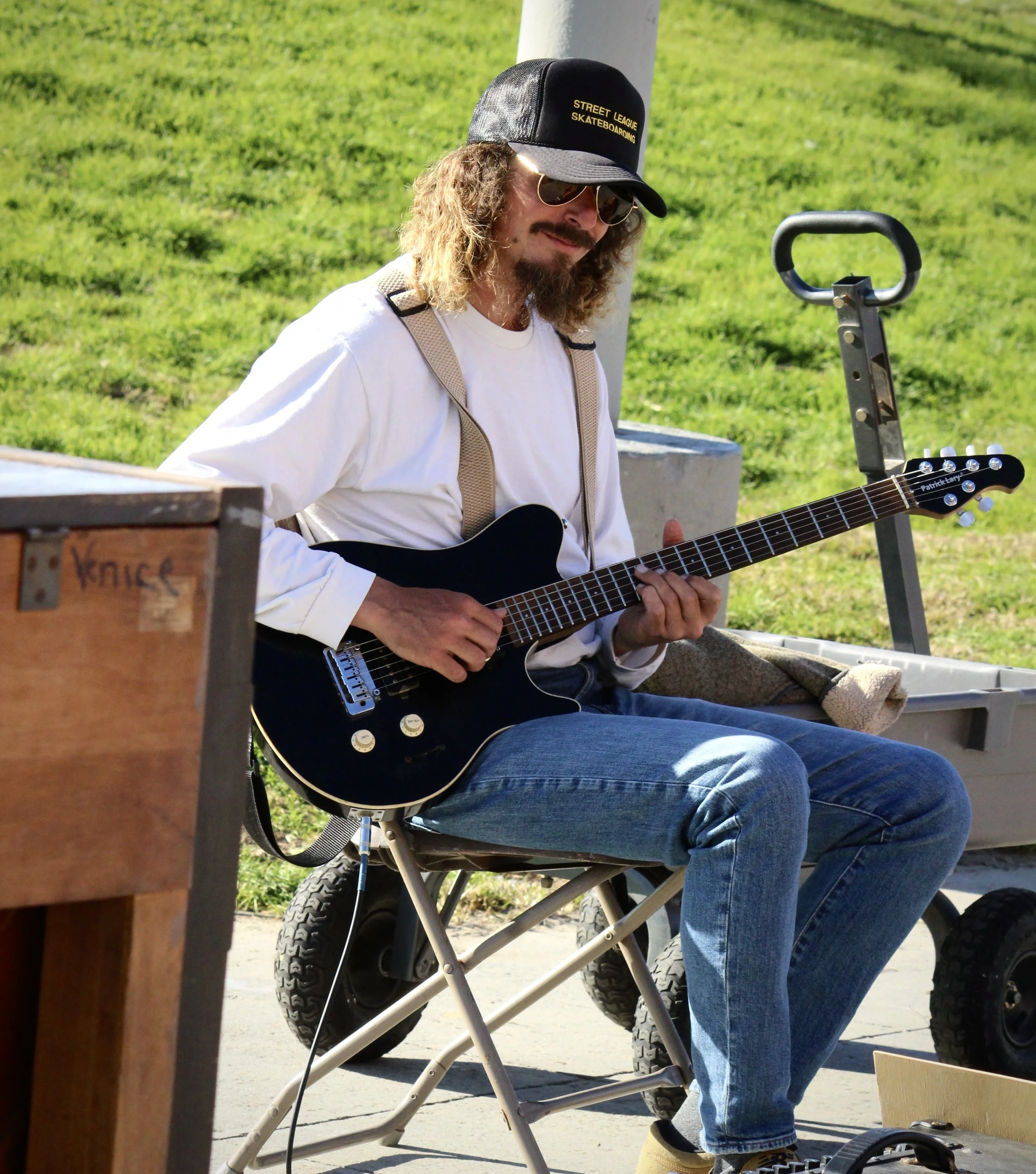Person sitting outdoors playing an electric guitar near a skateboarding ramp, wearing sunglasses, a cap, and casual clothes.
