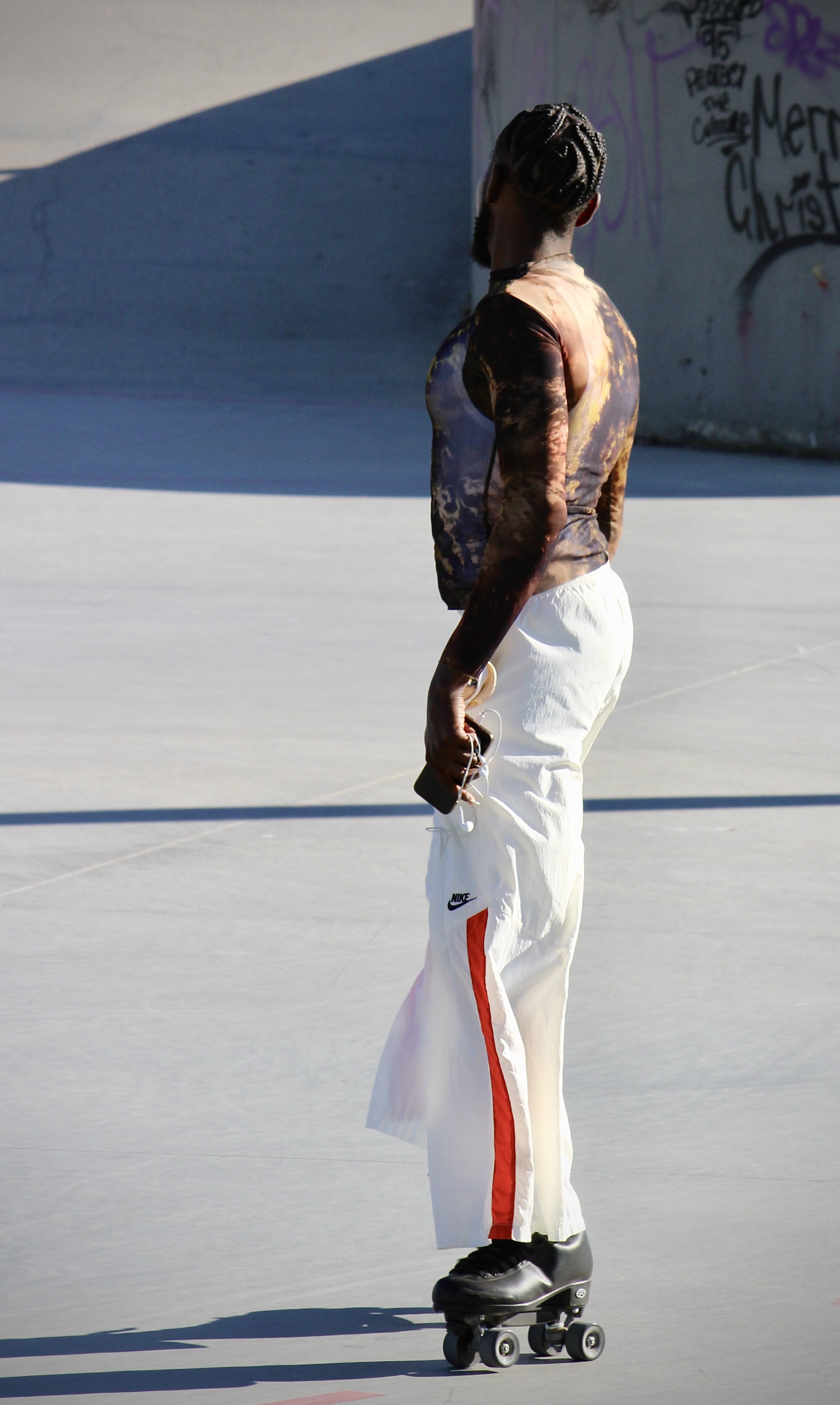 A person with braided hair skating on a concrete surface, wearing a patterned long-sleeve shirt and white pants with red and black stripes, holding a phone and earphones.