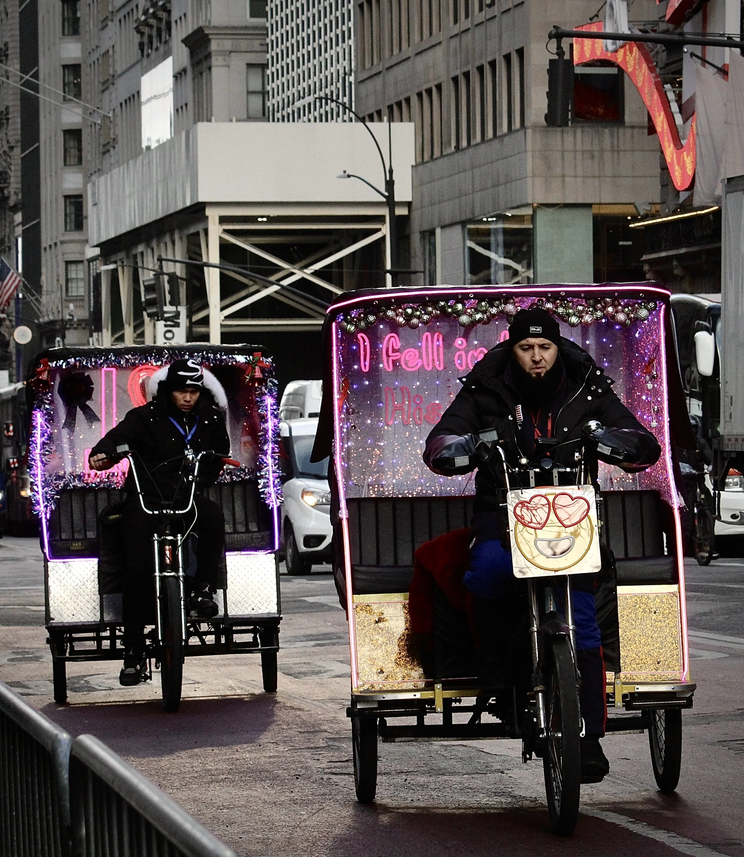 Two pedicabs decorated with colorful lights and Christmas ornaments, with one rider in a black jacket and knit cap pedaling on a city street with tall buildings in the background.