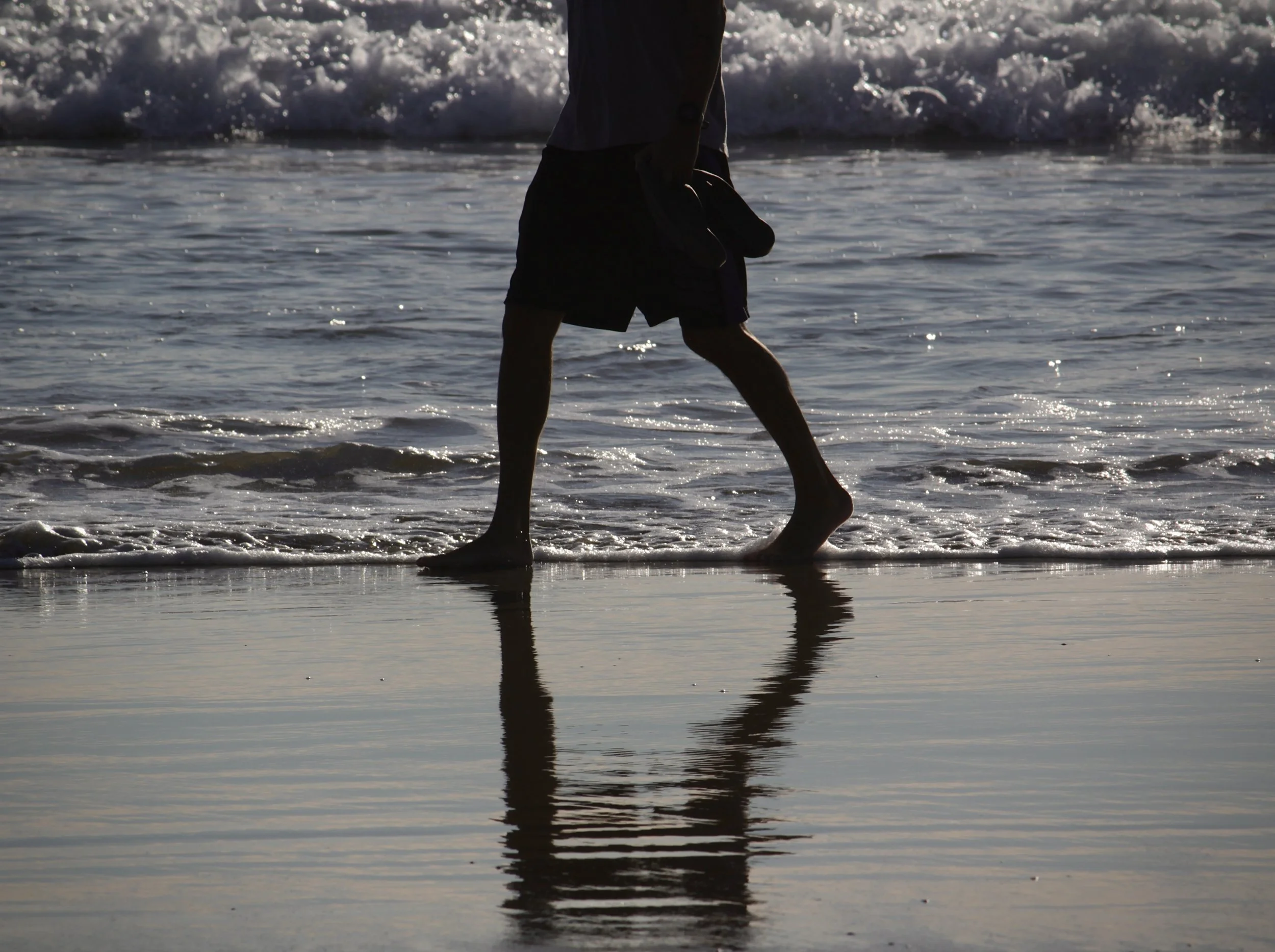 Person walking in the shallow water at the beach during sunset, with their reflection visible on the wet sand.