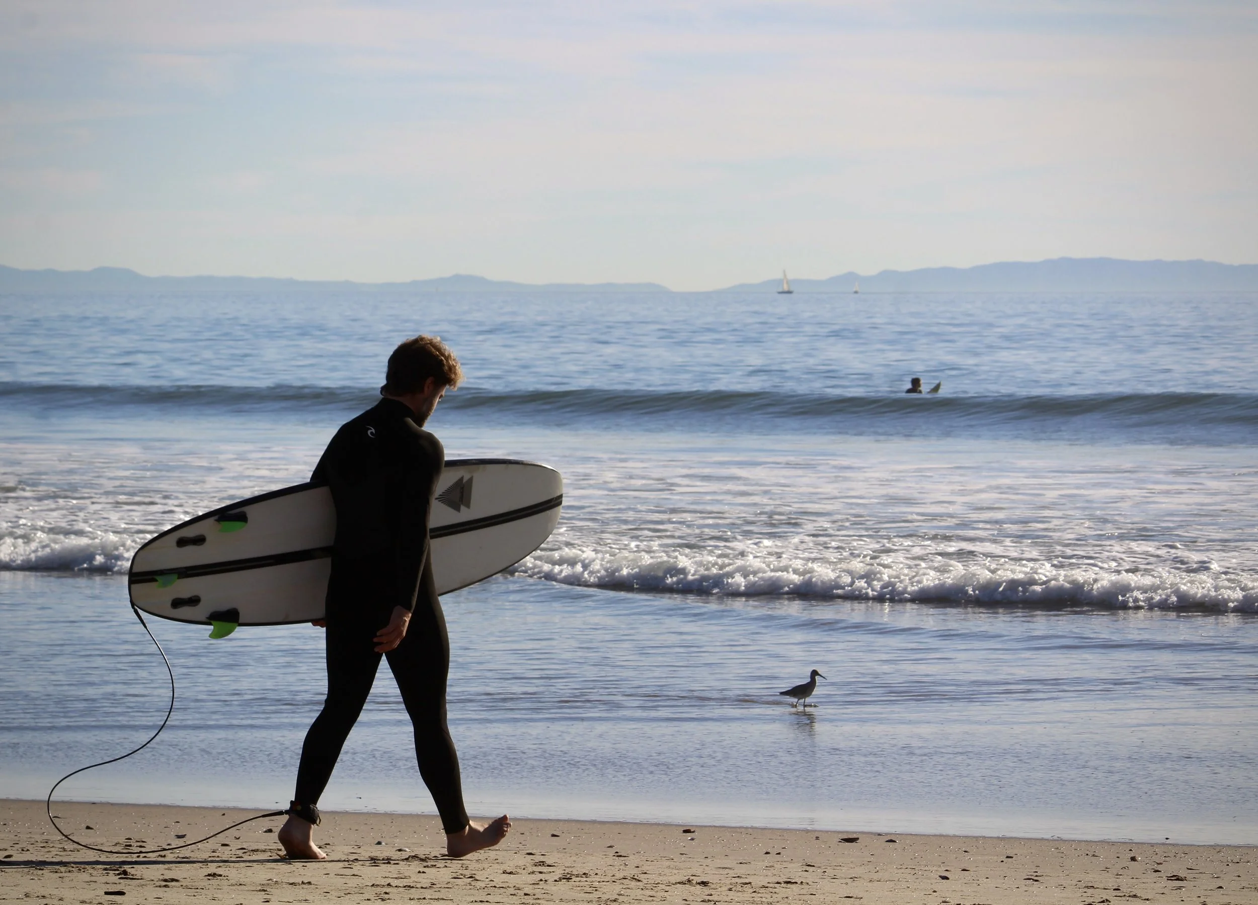 A person in black wetsuit walking on the beach with a white surfboard, holding it under their right arm. The person is barefoot, walking along the shoreline with gentle waves. In the background, a seagull is walking near the water, and a sailboat is 