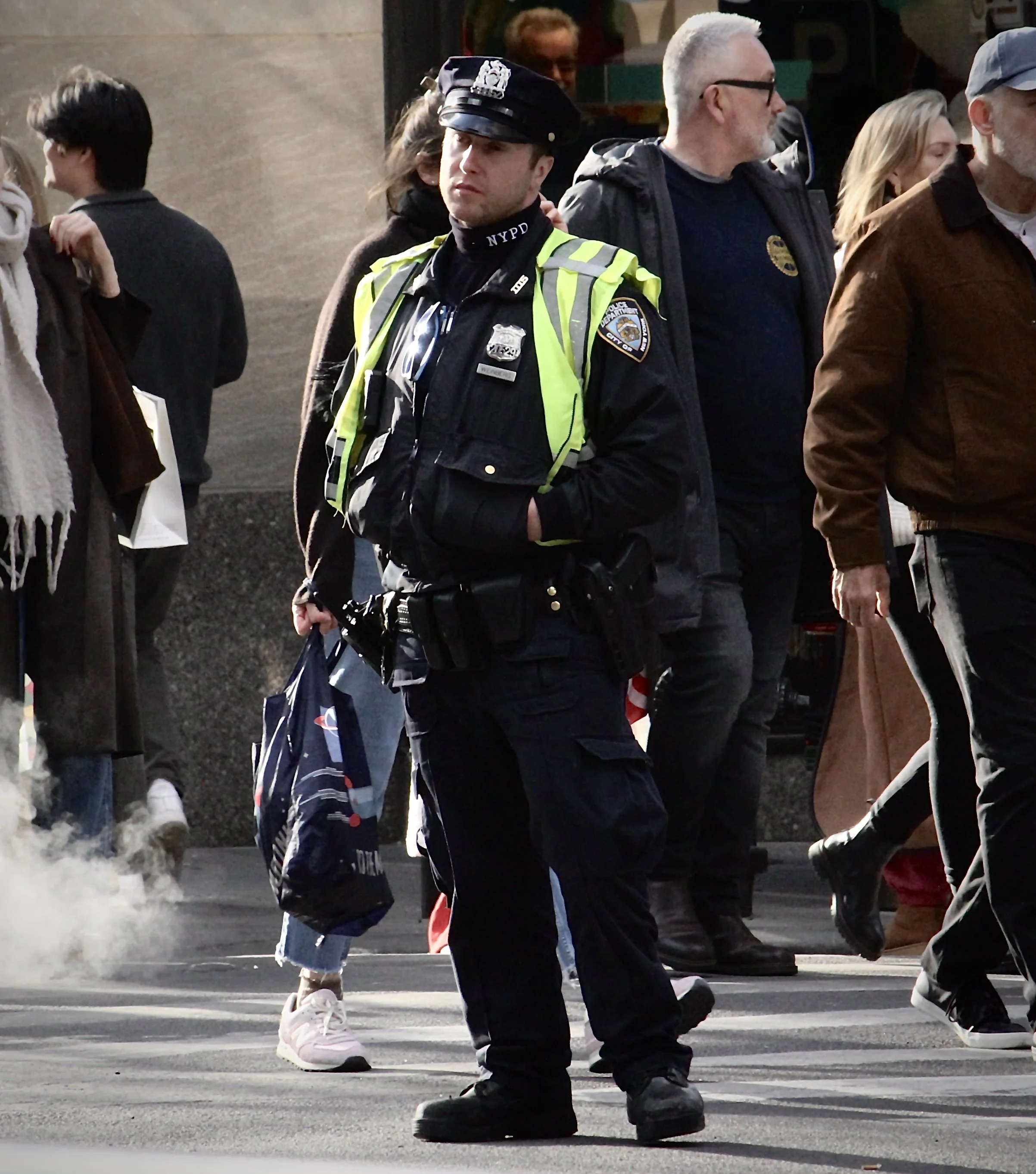 A police officer in a black uniform and yellow reflective vest standing at a crosswalk during daylight, with several pedestrians walking past him.