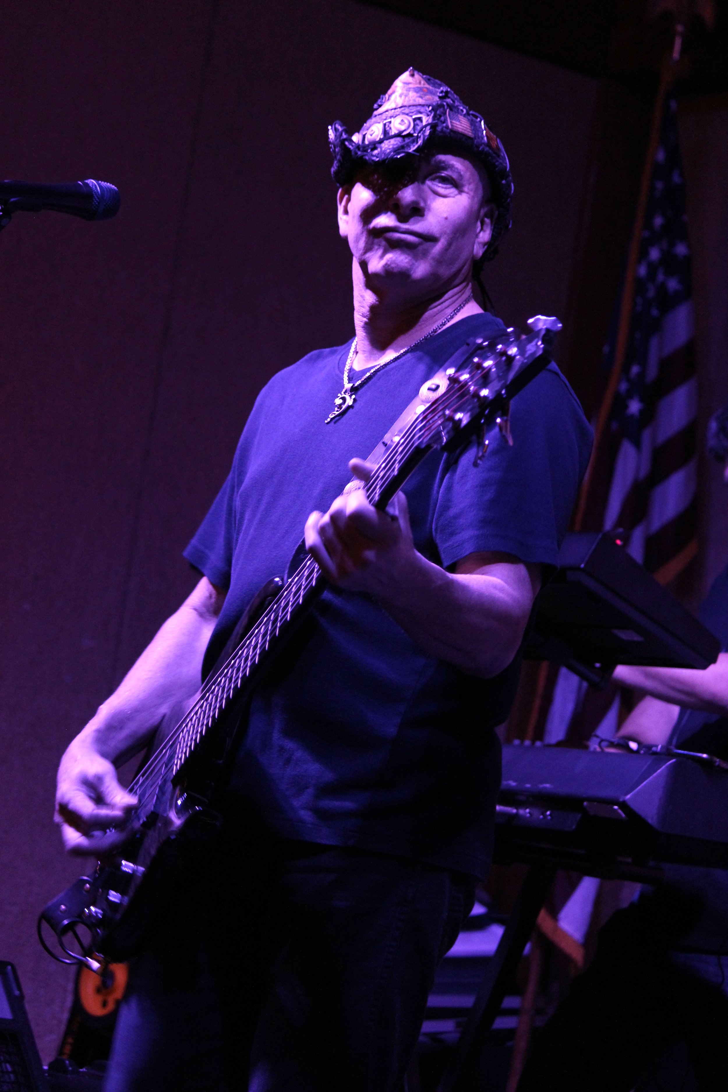A man wearing a decorative hat and a black t-shirt playing an electric guitar on stage with dark background, a microphone to his left, and American flag in the background.