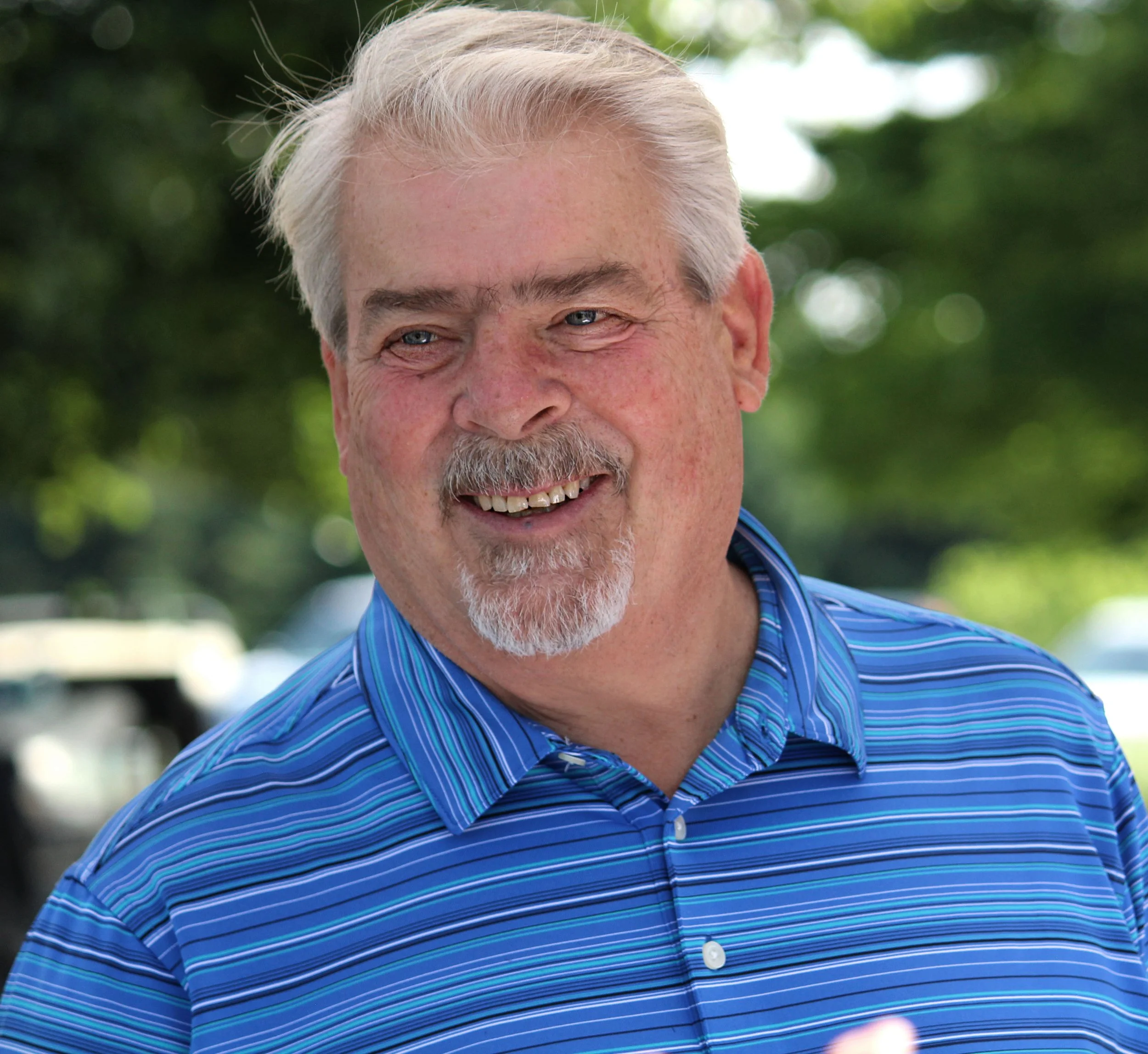 Close-up of smiling older man with grey hair and beard, wearing a blue striped shirt, outdoors with blurred green foliage in the background.