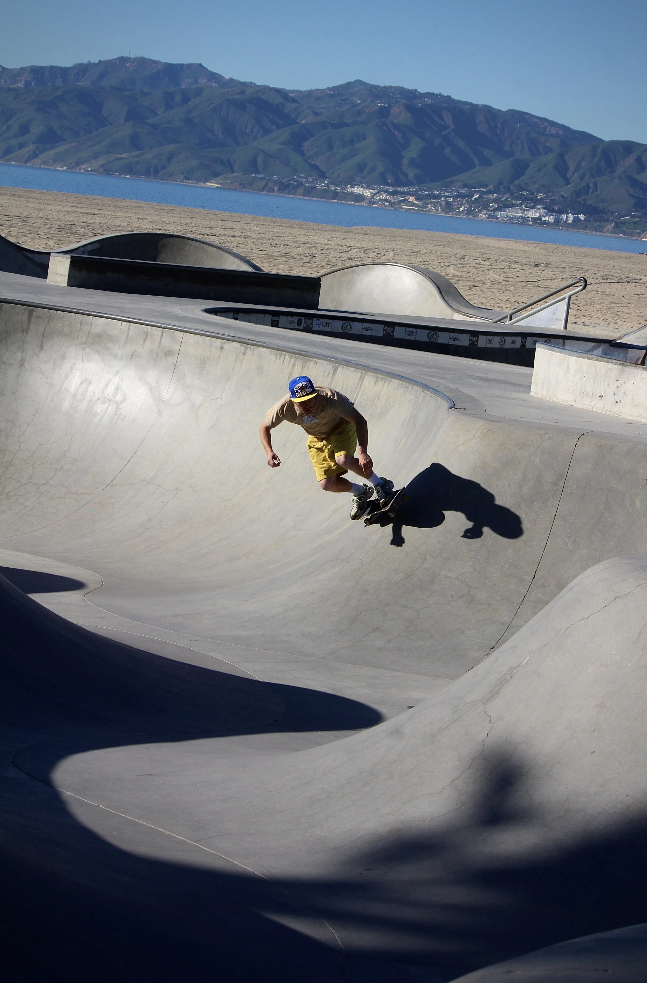 A skateboarder in yellow shorts and a blue cap skateboarding in a concrete skatepark with smooth curved bowls, with a landscape of mountains and water in the background.