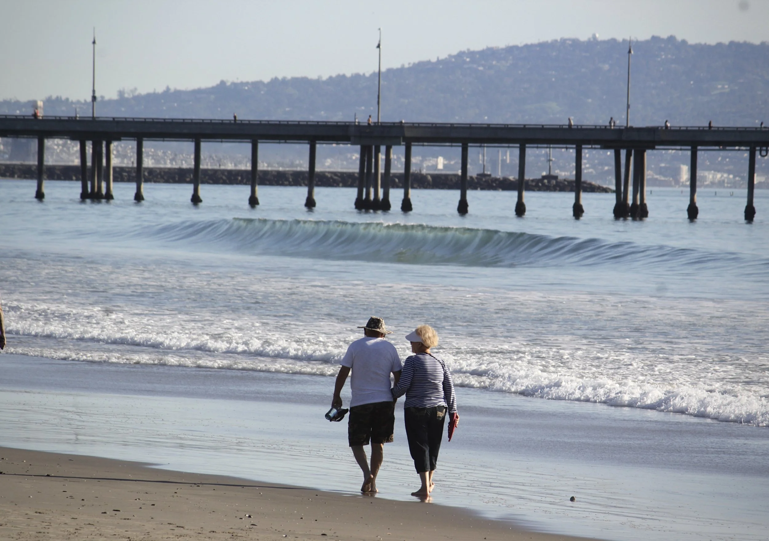 Two people walking along the beach near the water, with a pier in the background, during daylight.