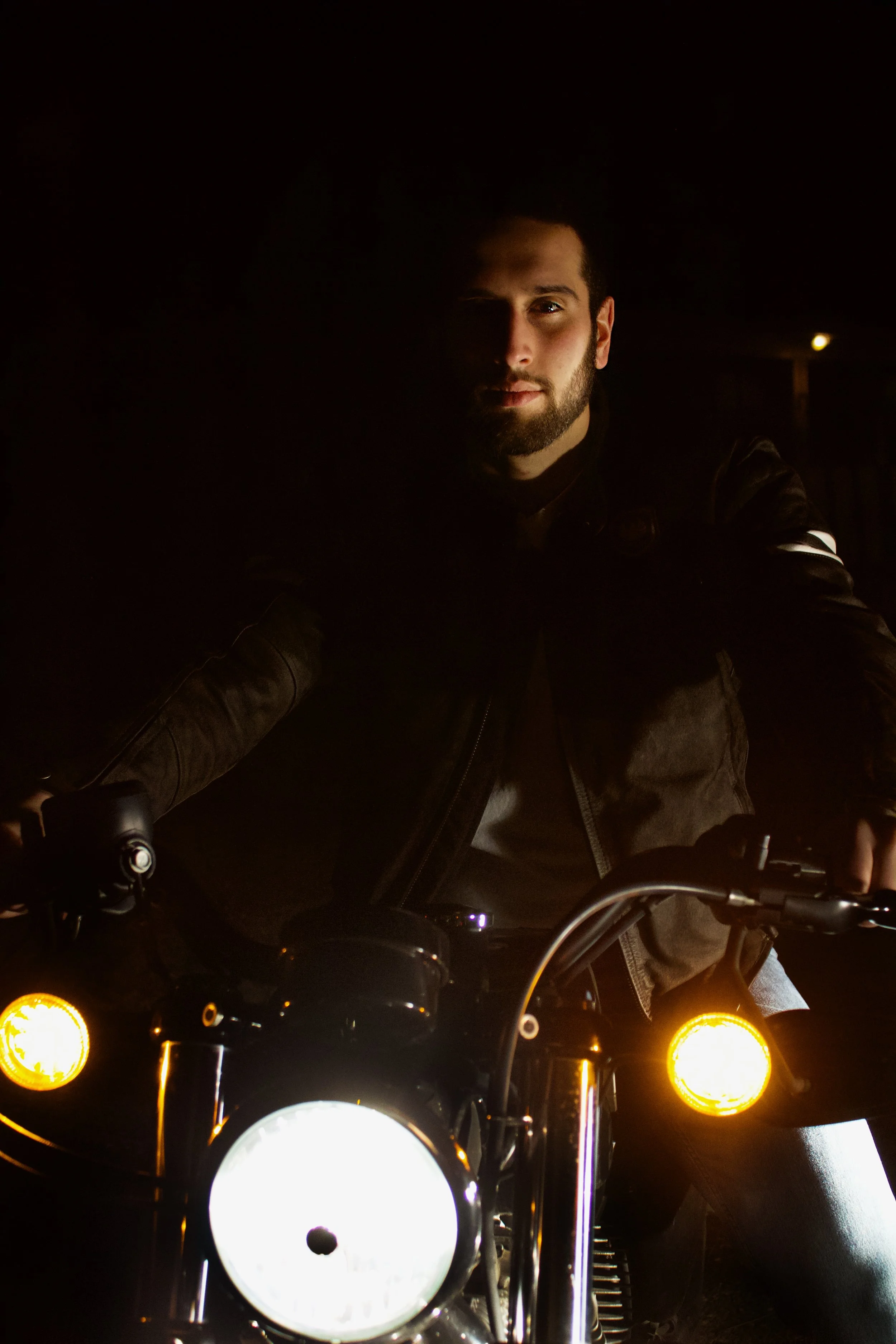 A man with a beard riding a motorcycle at night, illuminated by the motorcycle's headlights and turn signals.