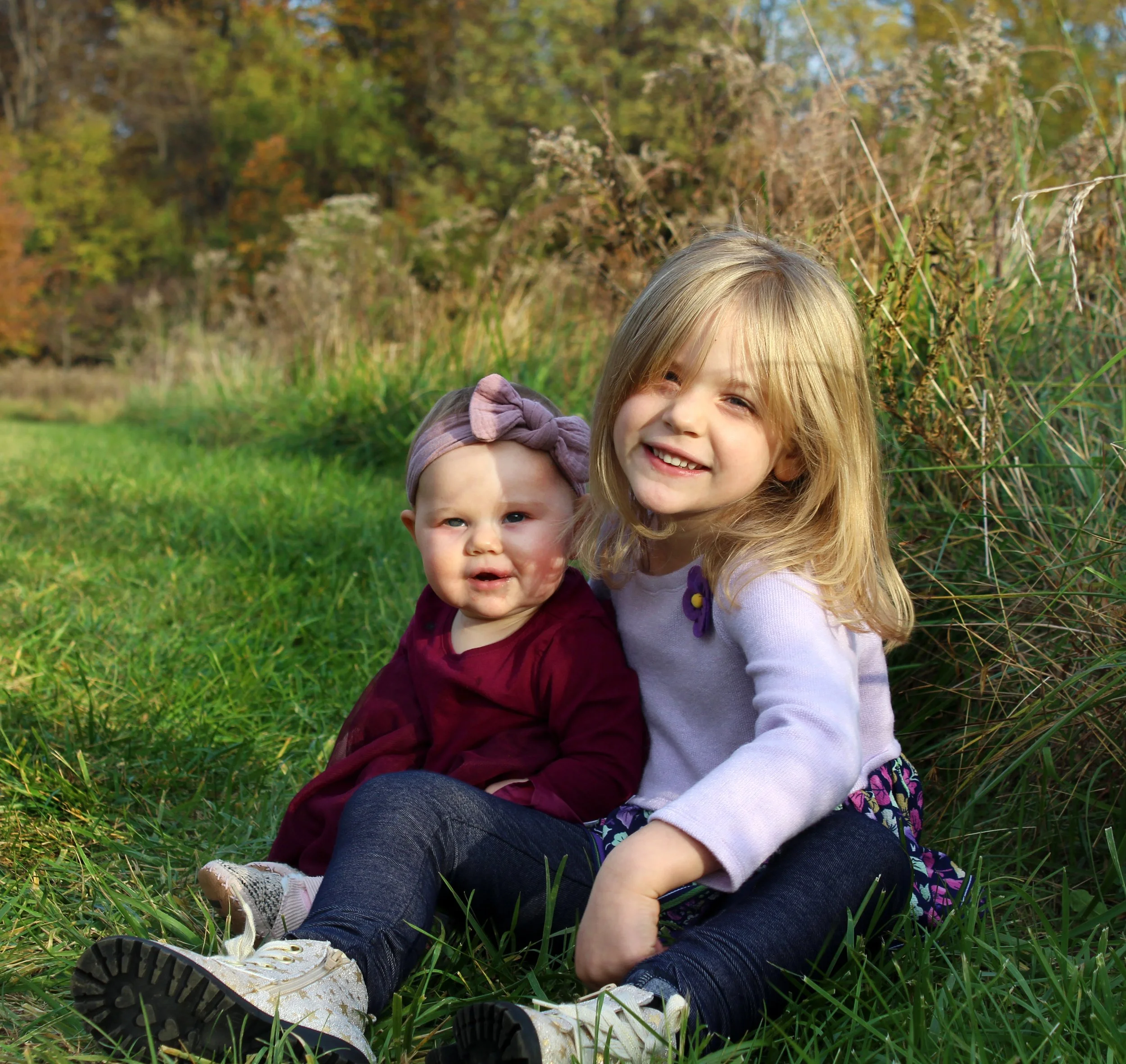 Two young girls sitting on grass outdoors, smiling, surrounded by tall grass and trees with autumn foliage.