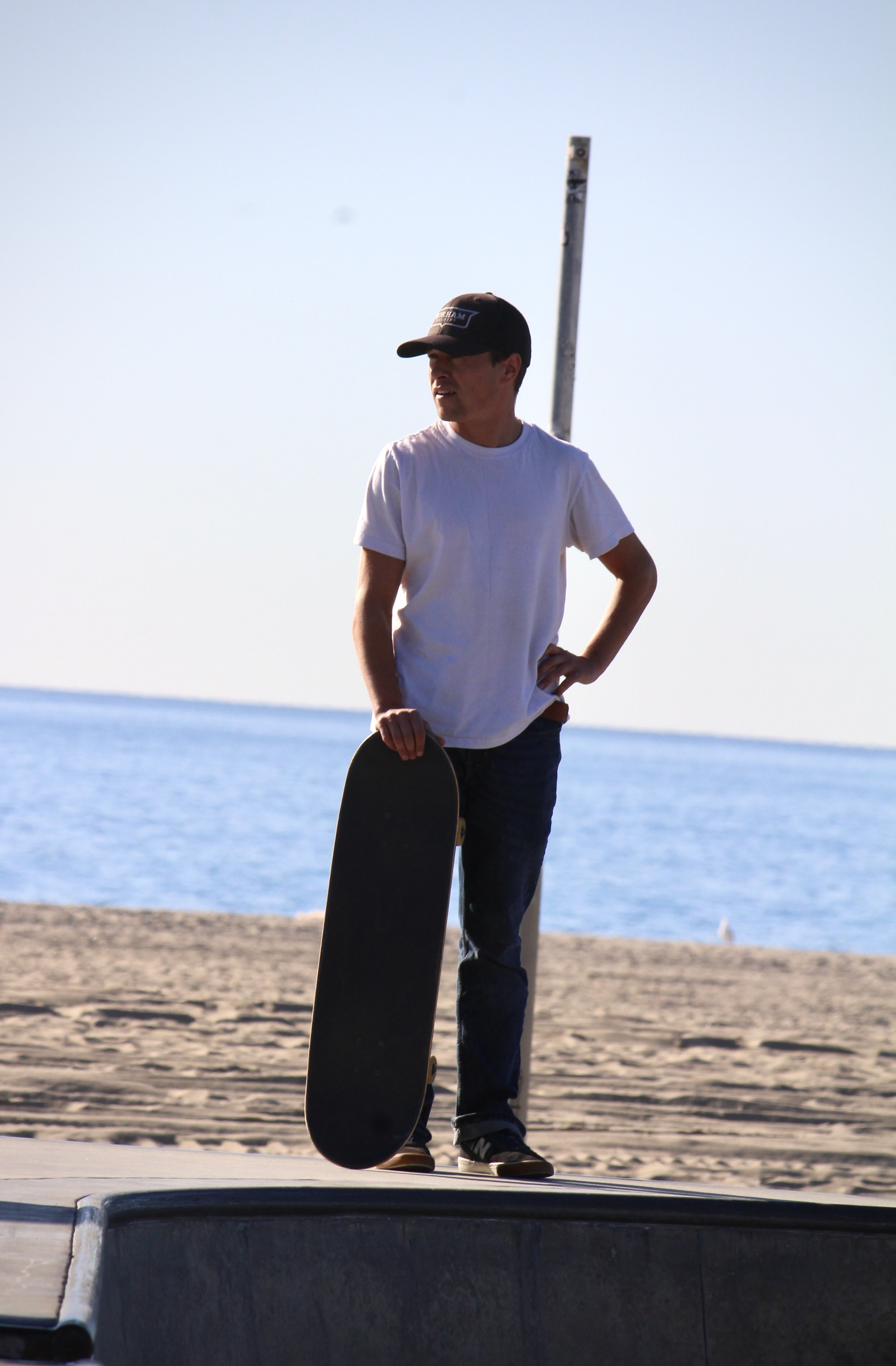 A man standing on a skateboard at a skate park near the beach, holding a skateboard in his right hand, wearing a baseball cap and white t-shirt, with the ocean and a pole in the background.