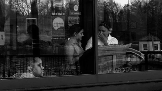 Three women sitting inside a restaurant or cafe, seen through a window, engaged in conversation.