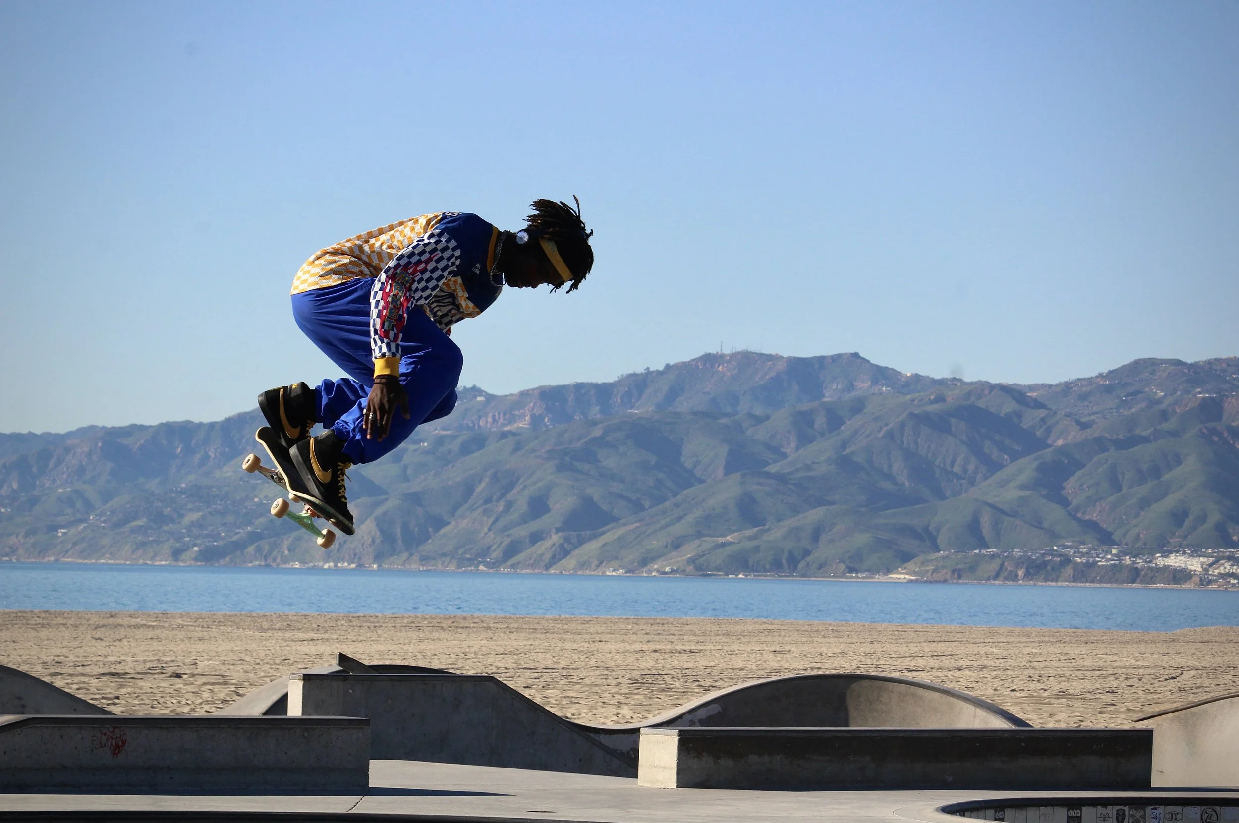 Skateboarder performing an aerial trick outdoors near a body of water with mountains in the background.