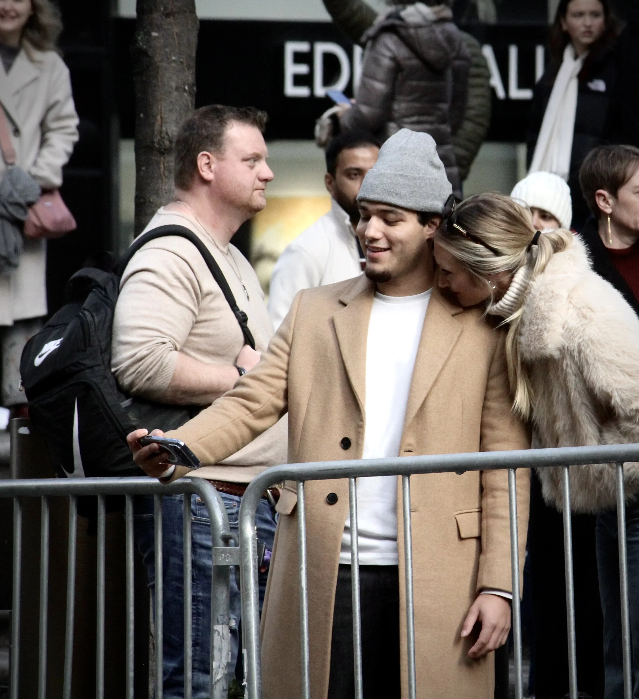 Group of people gathered behind a metal barricade, with a woman in a beige coat and gray beanie at the center, holding a phone, and others around her engaged in conversation or using their phones. There is a sign partially visible in the background.