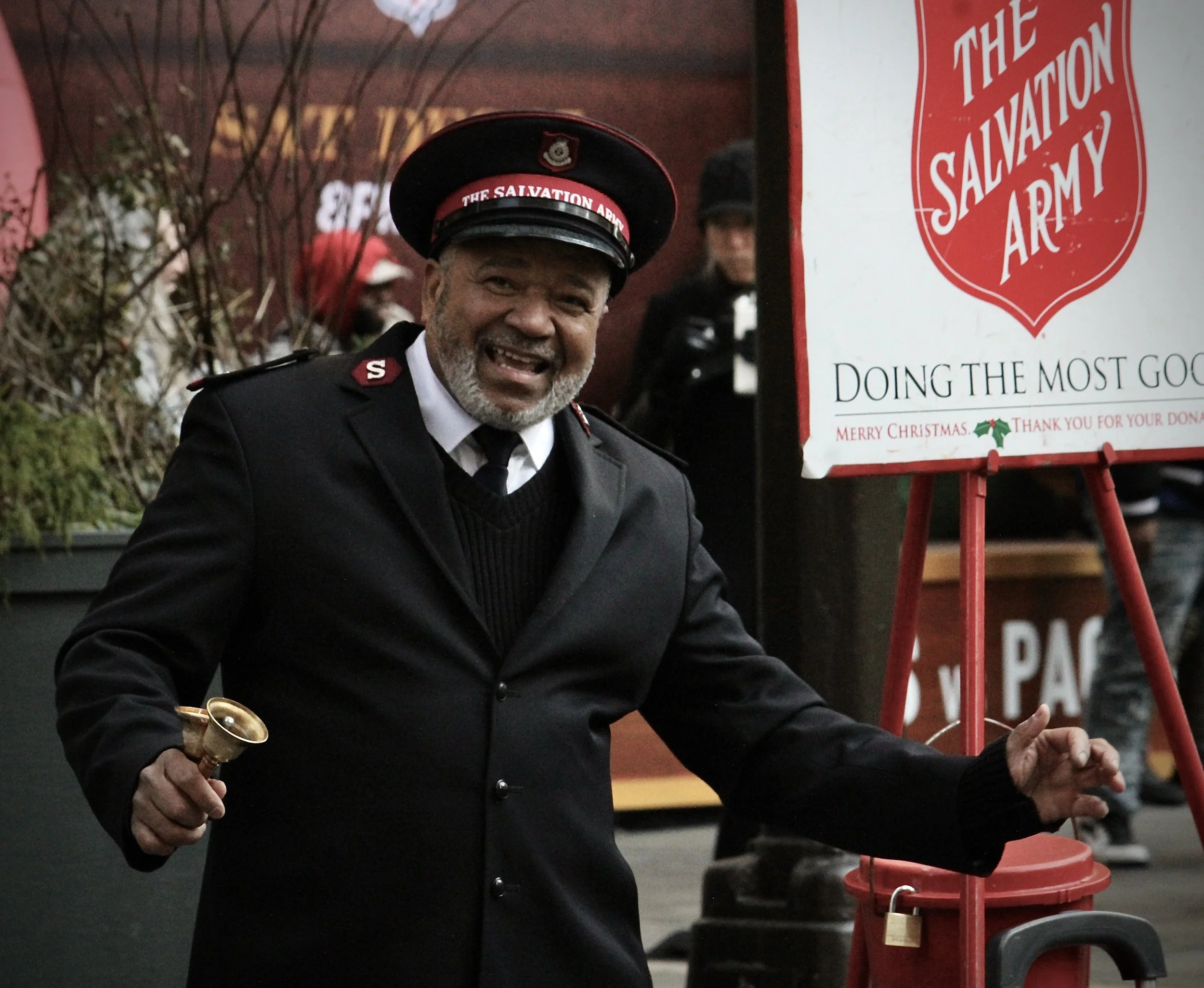 A man dressed in a Salvation Army uniform is smiling and holding a bell in his right hand, standing near a Salvation Army red donation kettle and stand during the Christmas season.