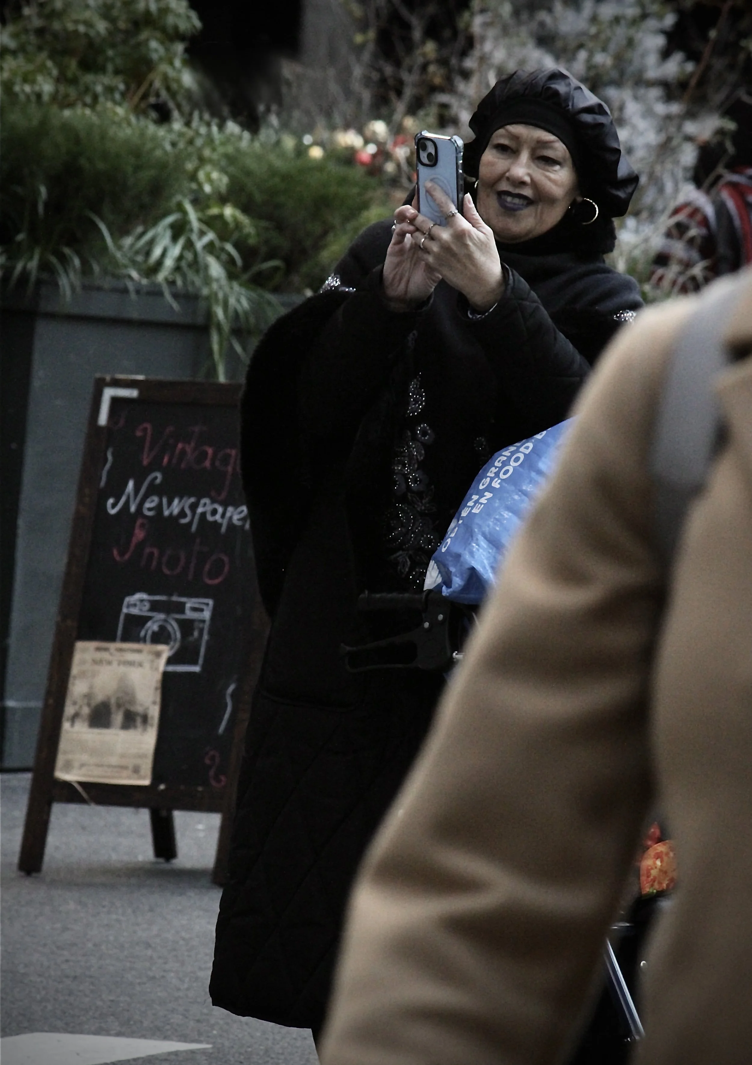A woman with dark lipstick and earrings taking a selfie with a smartphone at street-side in the evening, with a sign and plants in the background.