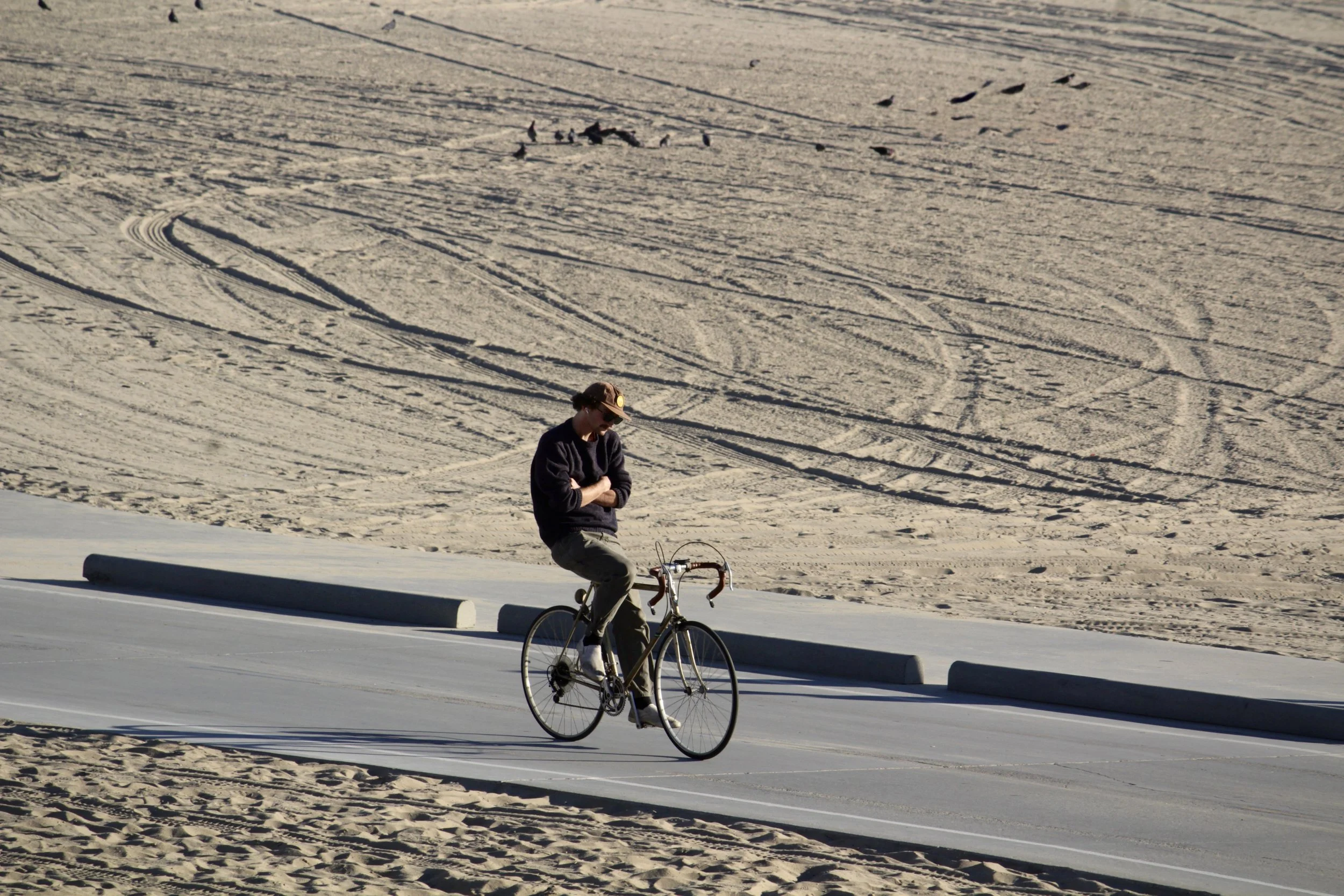 A person in a navy sweater, green pants, and a hat with sunglasses riding a bicycle along a paved road next to a sandy beach with tire marks.