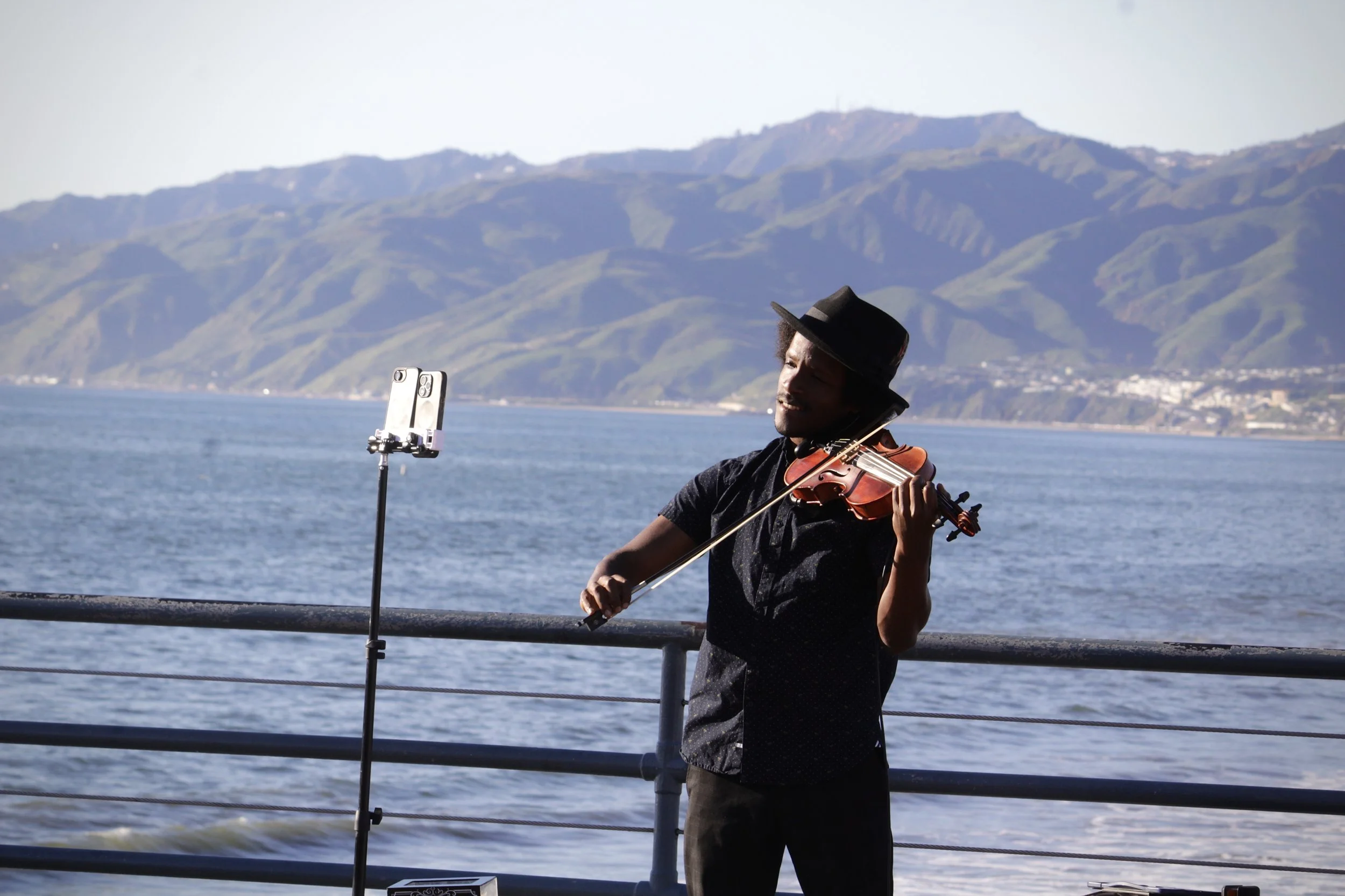 Man playing violin outdoors near water with mountains in the background, using a music stand.