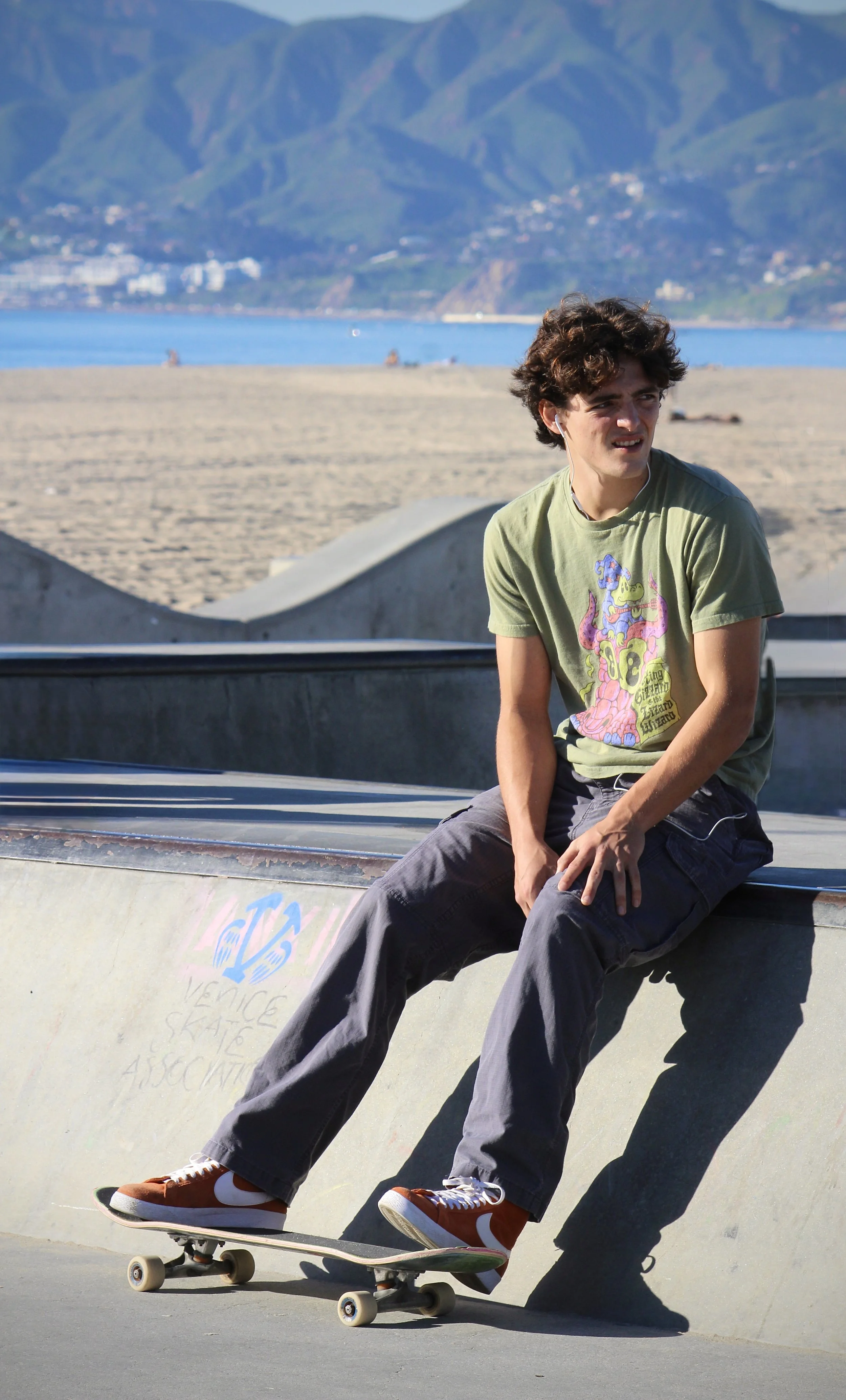 A young man skateboarding at a skate park near a beach, with mountains in the background.