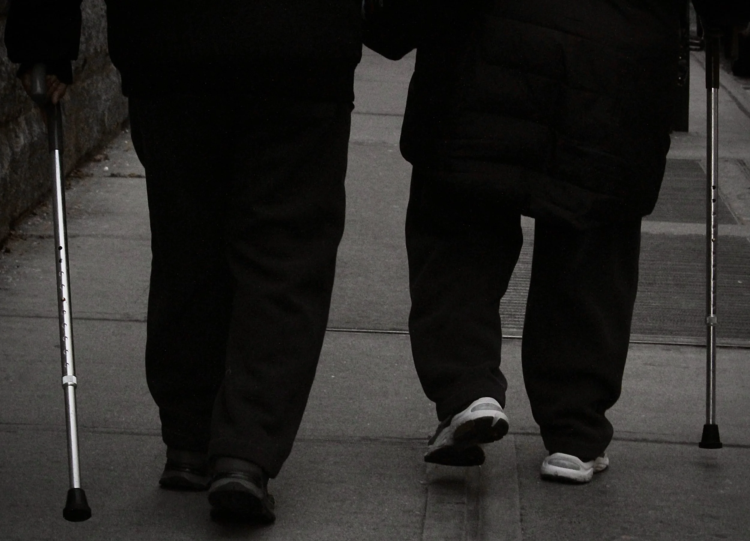 Two people walking with canes, wearing dark clothing and sneakers, walking side by side on a sidewalk.