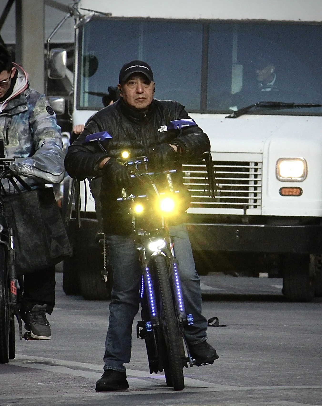 Man in black jacket and cap sitting on a black electric bike with bright yellow headlights, in front of a white truck, with other people and vehicles nearby.