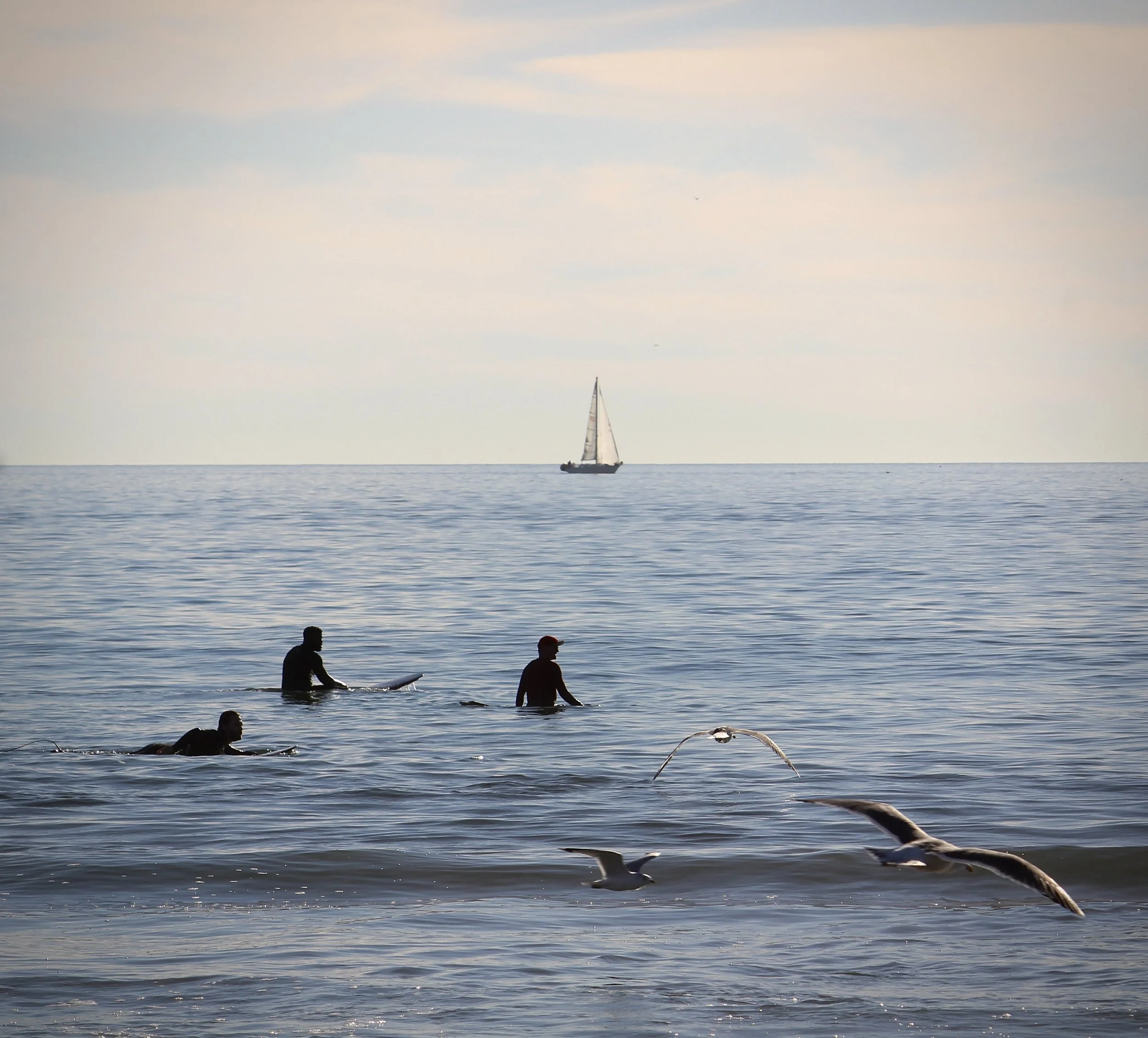 Sailboat on calm ocean with people swimming and a seagull flying overhead under a partly cloudy sky.