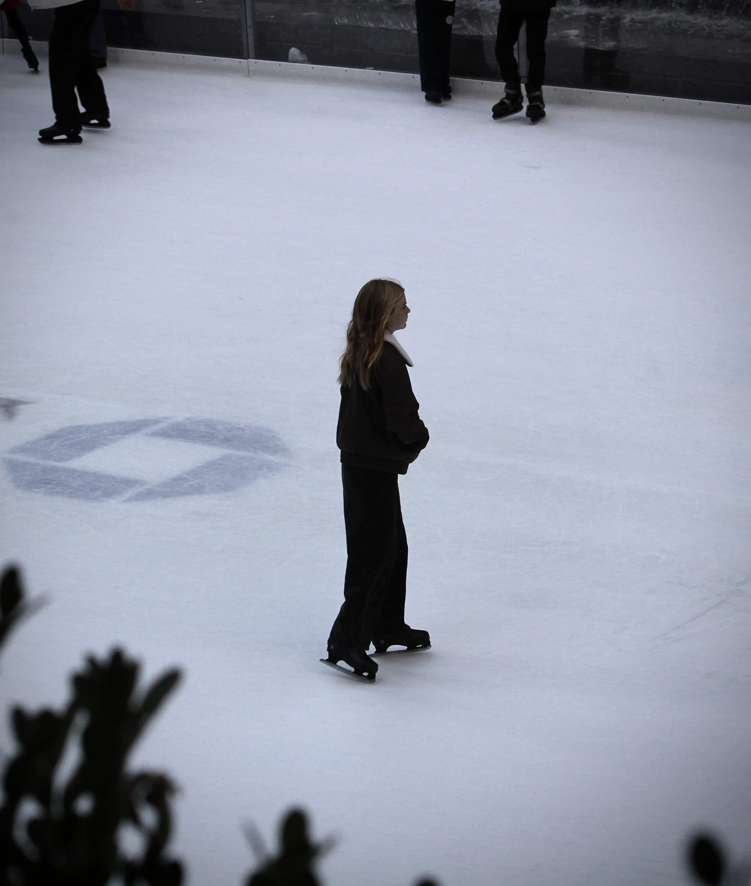 A person ice skating on an ice rink, seen from the side with their profile visible, wearing a dark jacket and pants, and ice skates. There are other people standing in the background near a barrier, and some blurred shapes in the foreground.