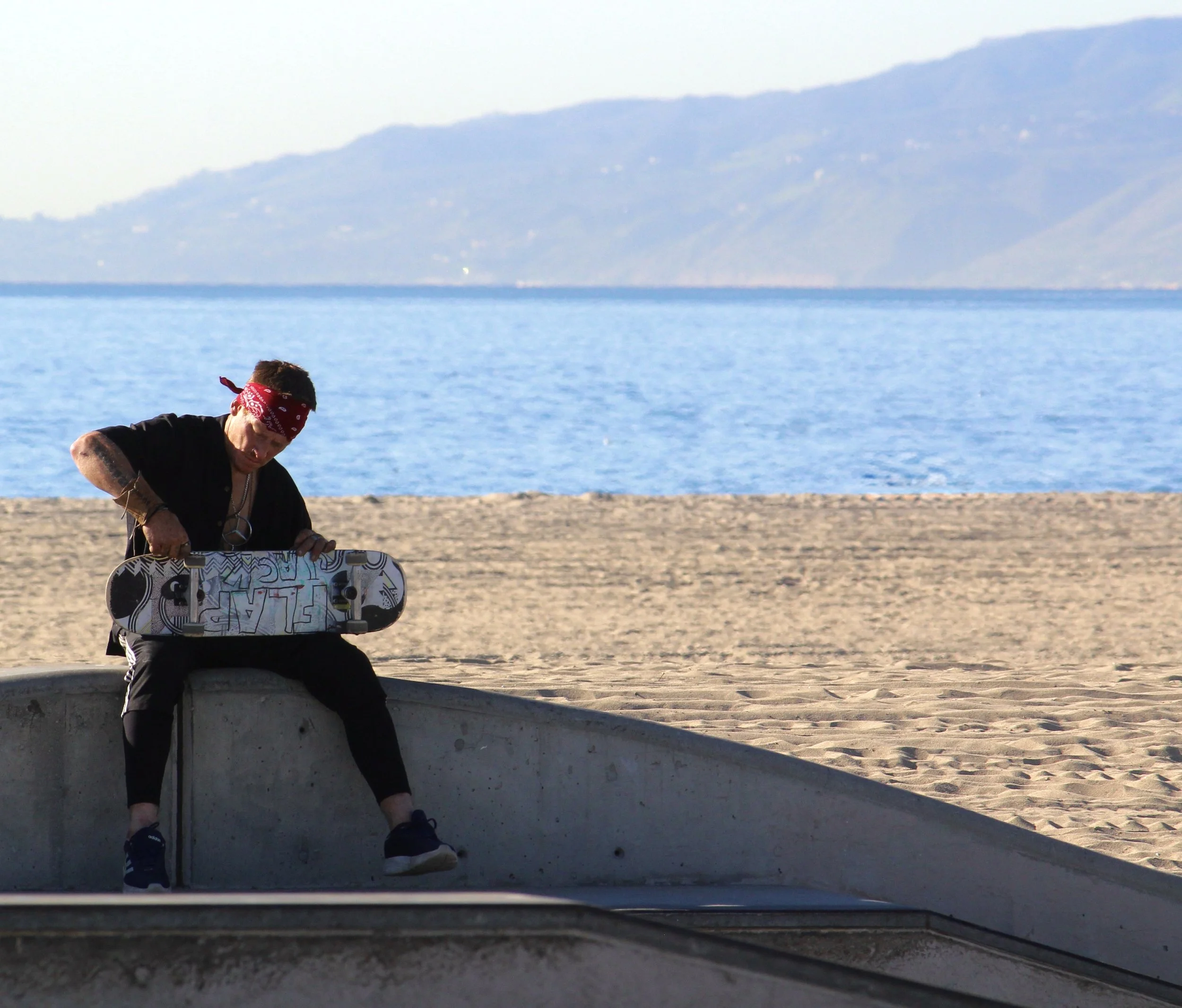 A person with a red bandana on their head sits on a ledge at the beach, holding a skateboard with a colorful design, with the ocean and mountains in the background.