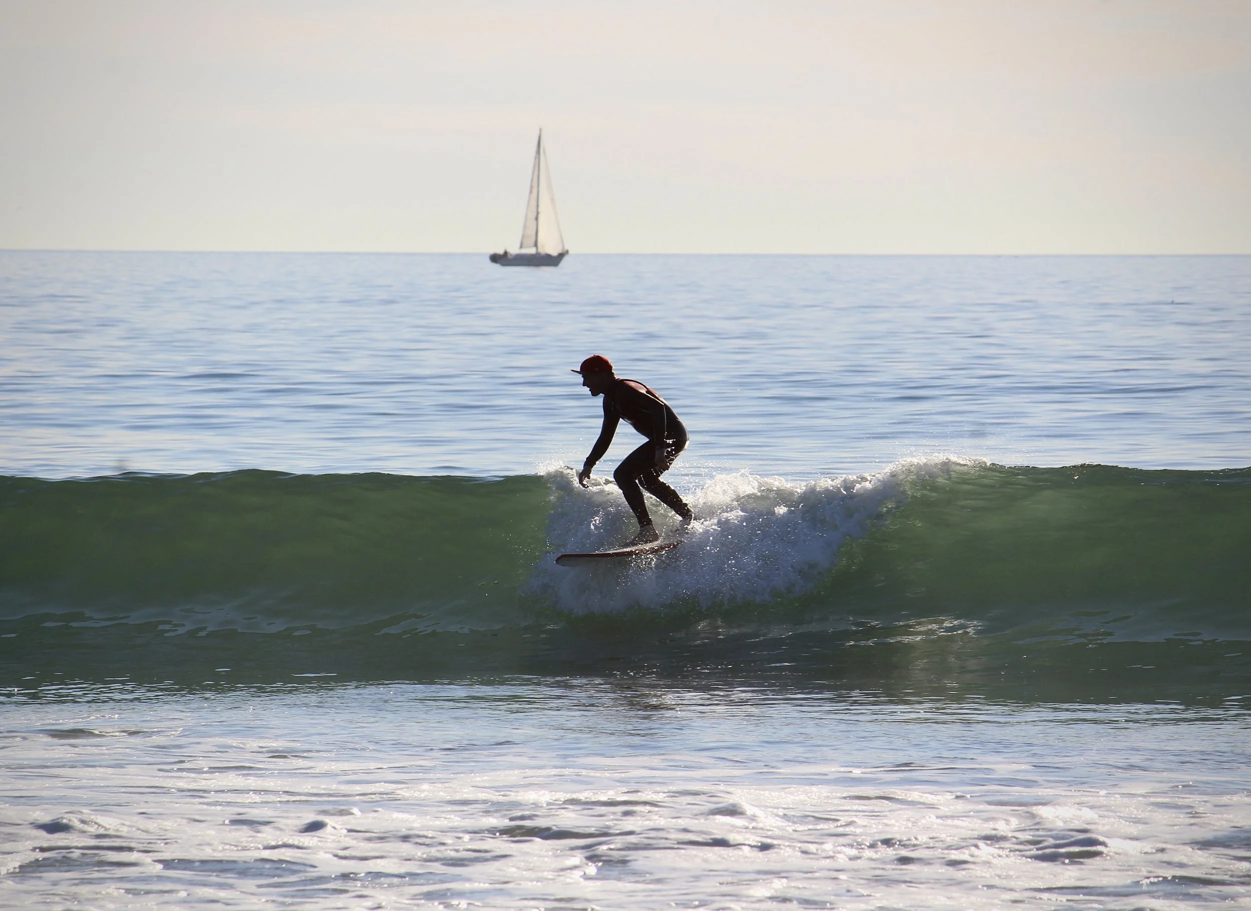 A person wearing a red helmet and black wetsuit riding a surfboard on a small wave in the ocean with a sailboat in the background.