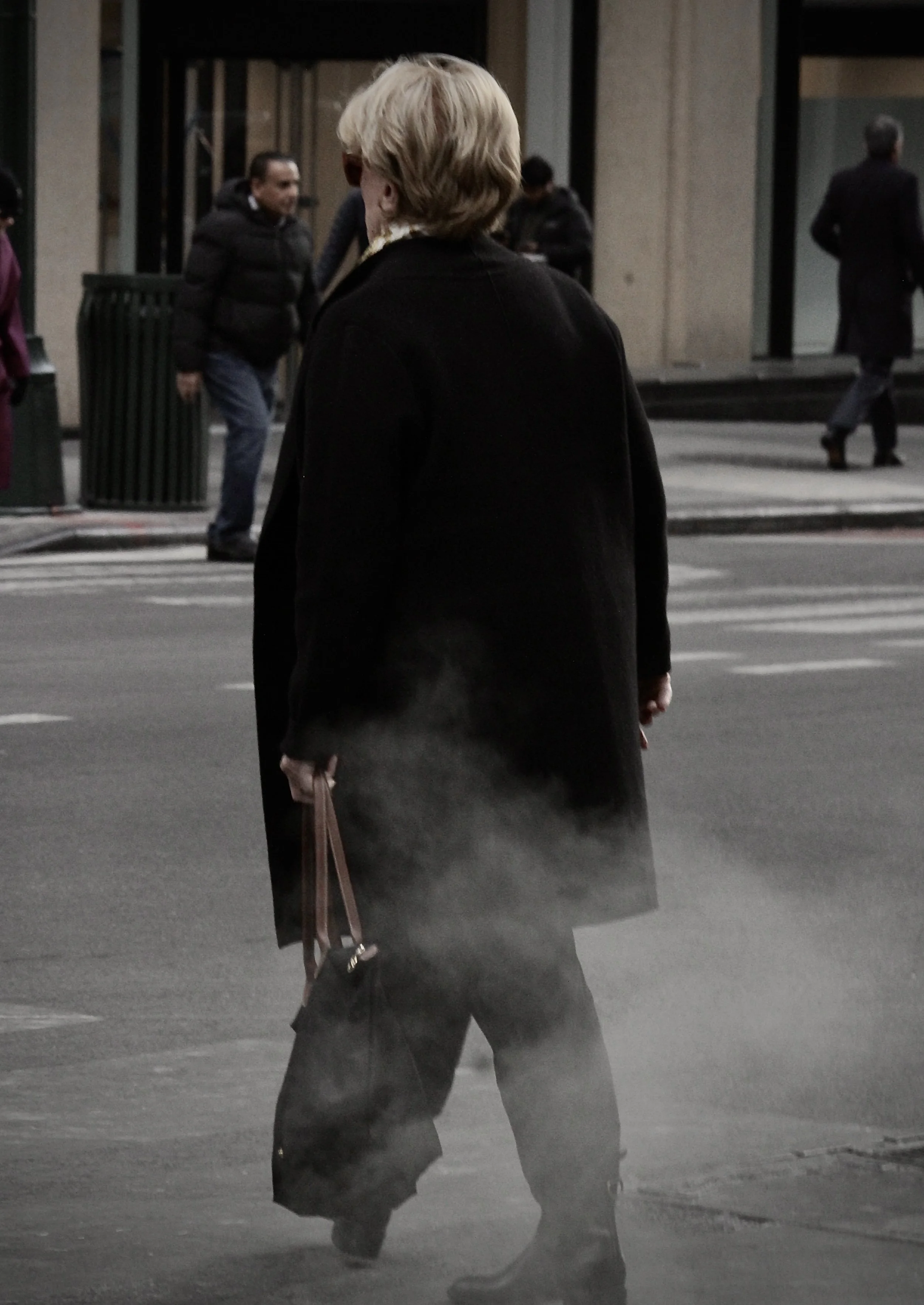 A woman wearing a black coat and pants, holding a brown handbag, walks on a city street at night, with mist or steam near her feet.