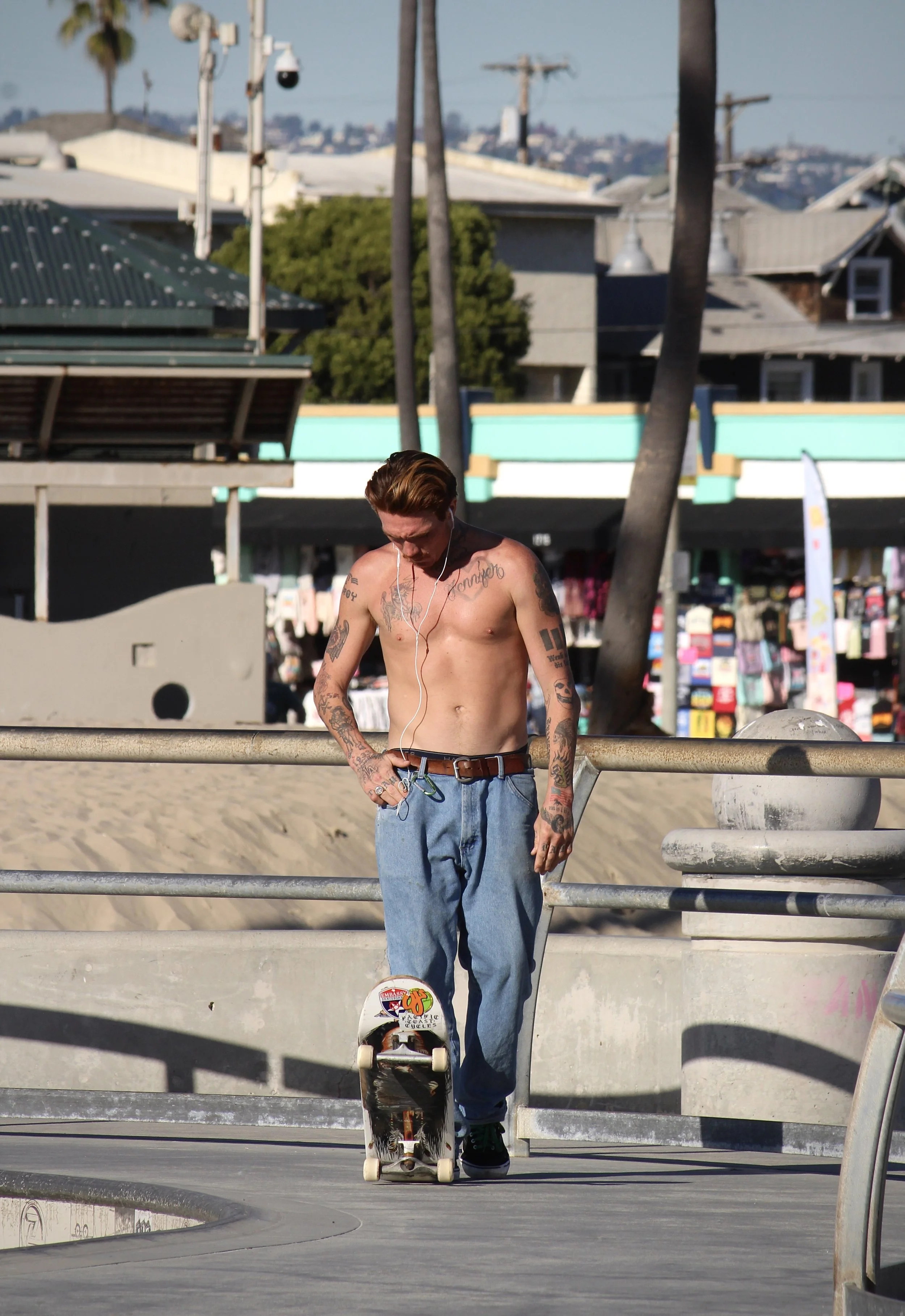 A shirtless male skateboarder with tattoos, wearing blue jeans and black sneakers, standing near a skate park railing, looking down at his skateboard with a colorful sticker, during daylight.