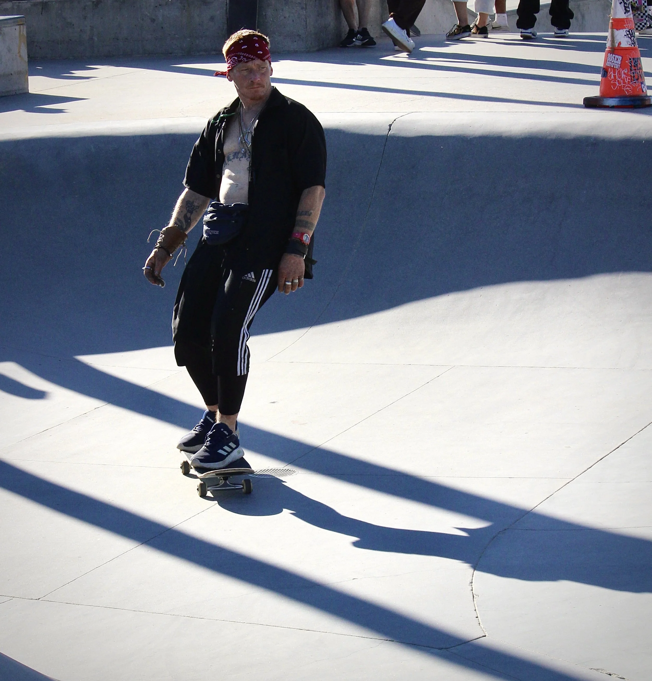 A man with a red bandana, tattoos, and wearing black Adidas pants skateboarding outdoors on a sunny day, with shadows cast on the concrete ground and several people walking in the background.