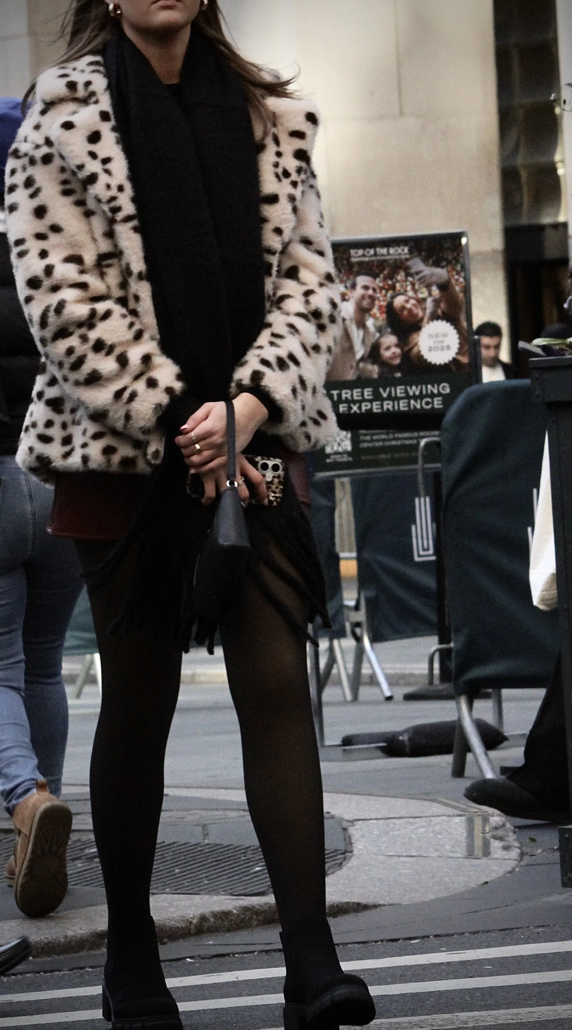 A woman wearing a leopard print coat, black tights, black shoes, and a black scarf, standing on a city street during the daytime.