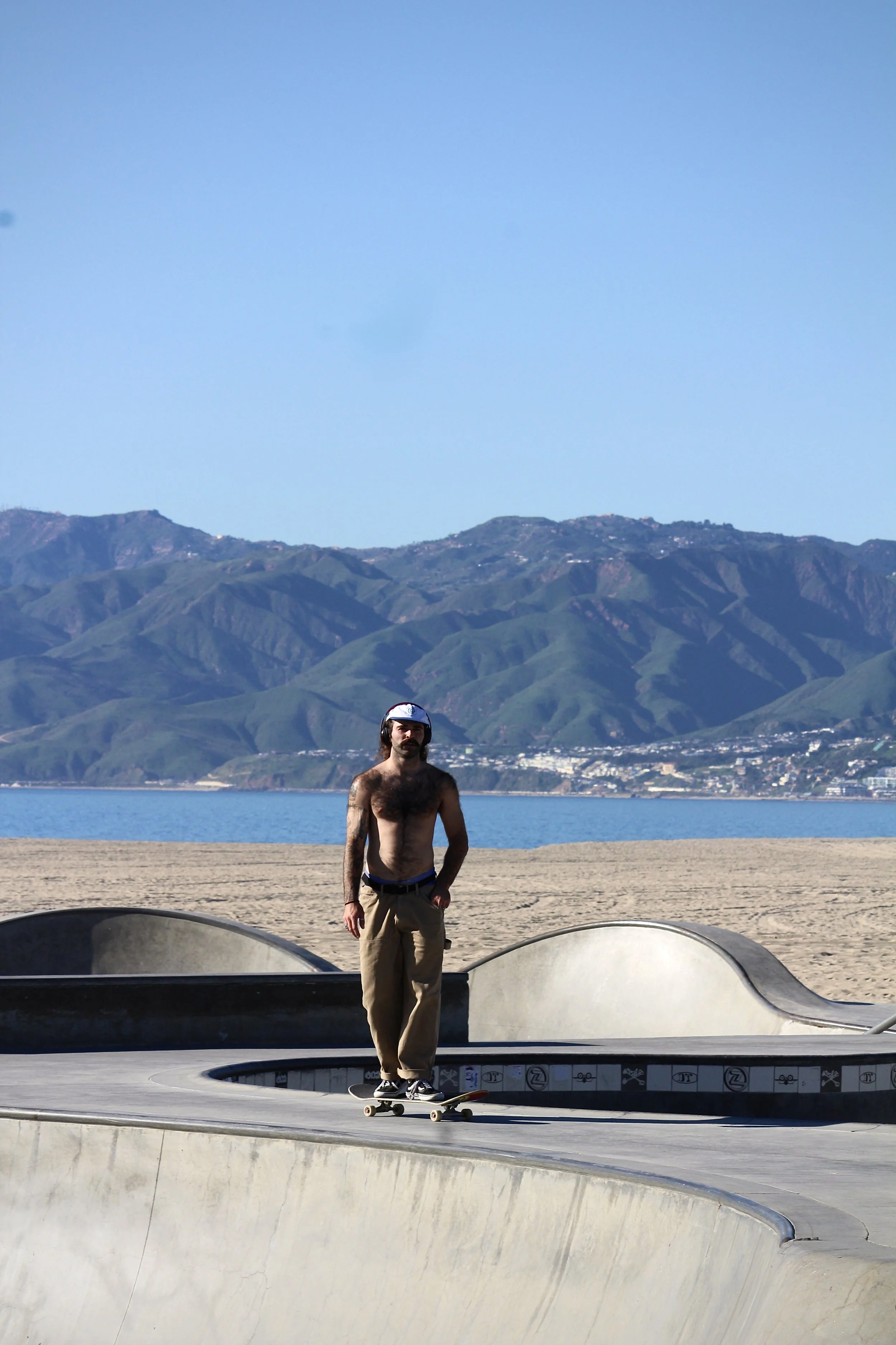 A shirtless man with tattoos wearing a helmet skateboarding at a skate park with green hills and mountains in the background.